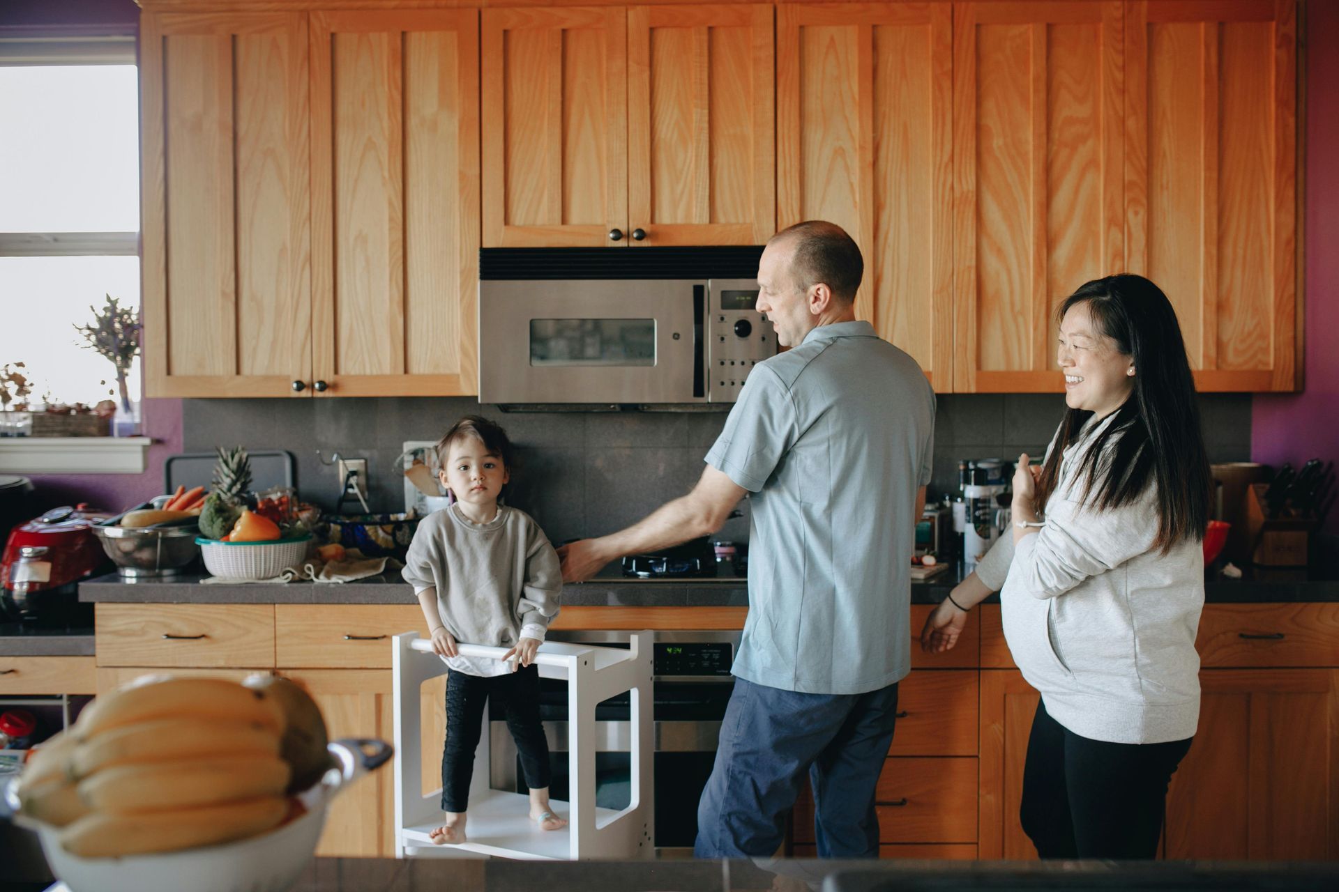 Family in kitchen: Child on step, parents cooking. Wooden cabinets, purple wall, fruits on counter, sunny.