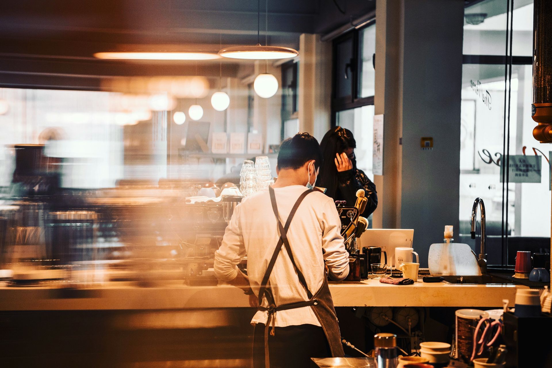 A barista wearing an apron works at a cafe counter, with a blurred view of customers behind him.