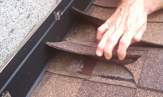 Hand inspecting damaged roof shingles near the gutter on a brown shingled roof