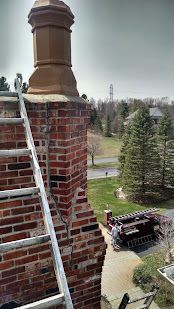 Brick chimney with a ladder on a house roof, overlooking trees and a driveway below.