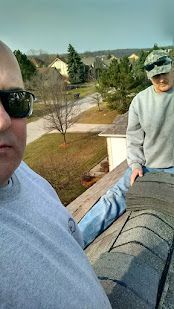 Two people on a rooftop, one taking a selfie, with a suburban neighborhood in the background