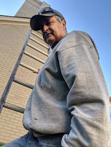 Man in a gray sweatshirt and cap standing beside a brick building and ladder, viewed from below
