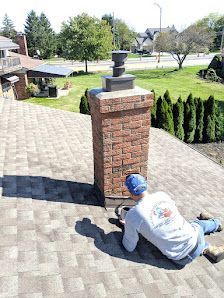 Worker repairing brick pillar on a paved driveway in a sunny residential yard.
