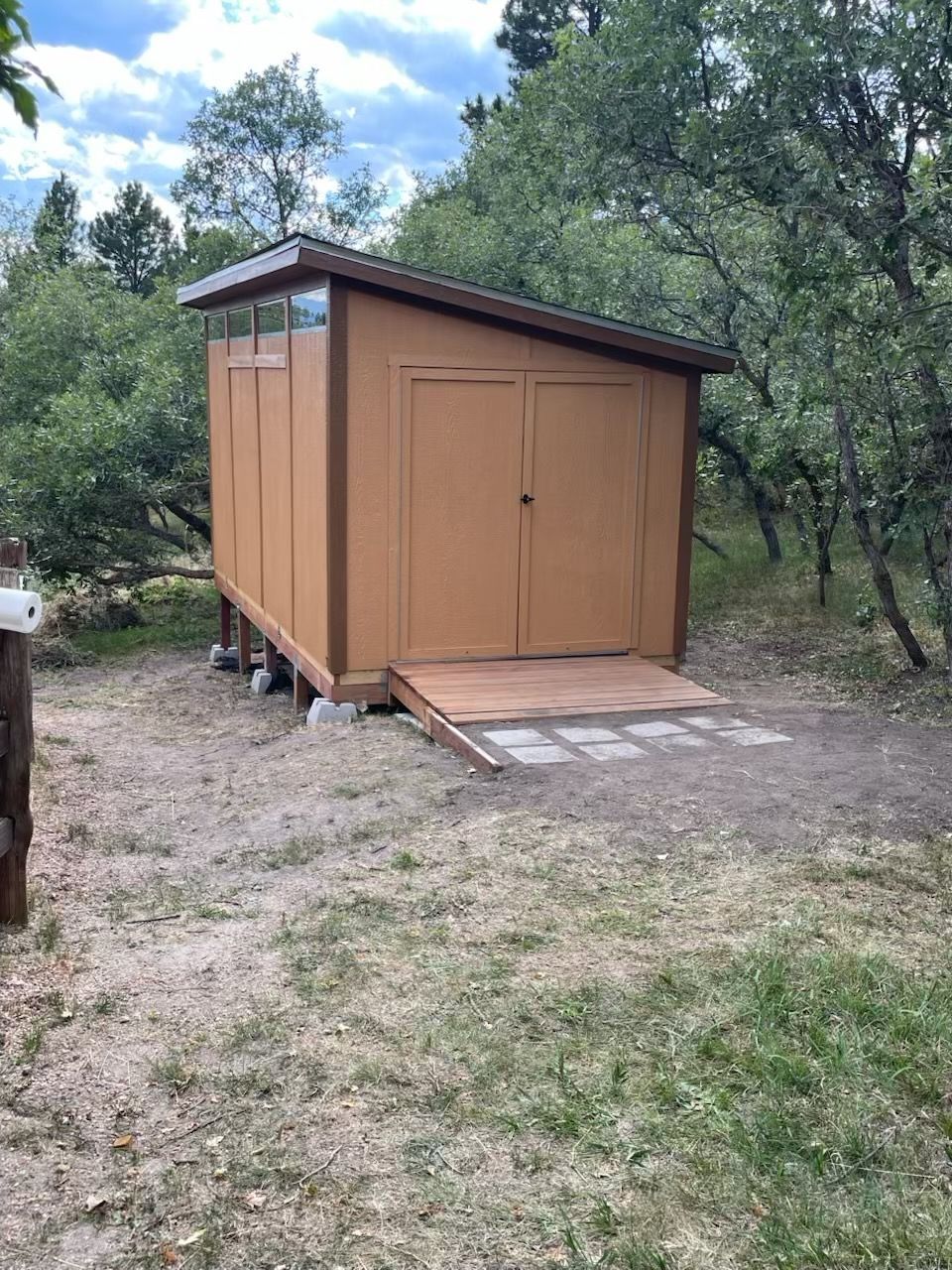 Small brown wooden shed with double doors and ramp in a grassy yard surrounded by trees