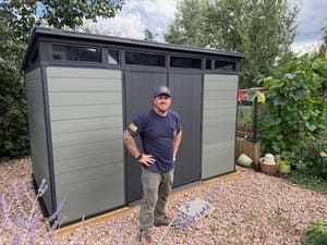 Man standing in front of a gray garden shed in a gravel yard with greenery and trees around it.