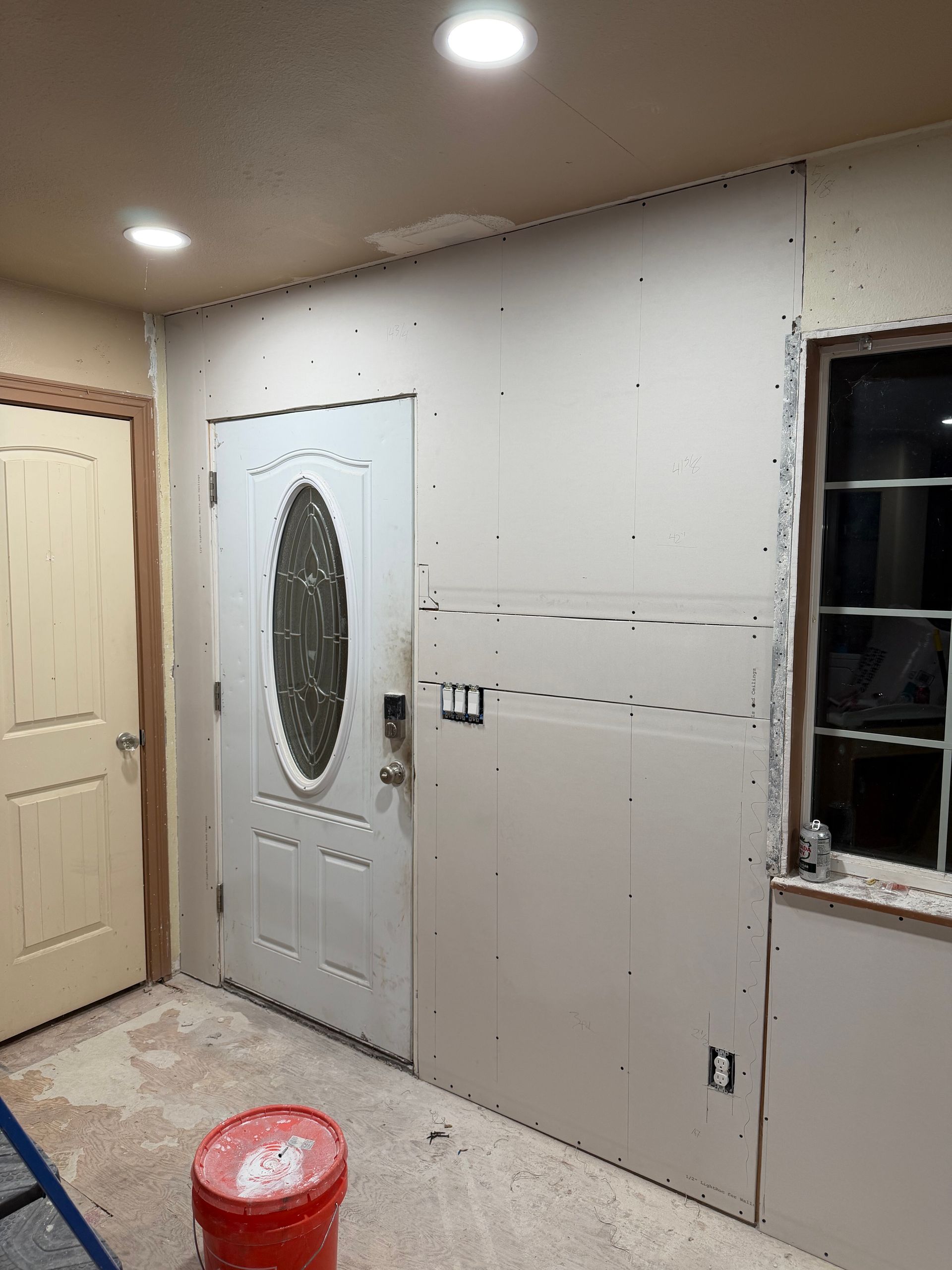 Unfinished room with drywall, a white door, window opening, and a red bucket on the floor.