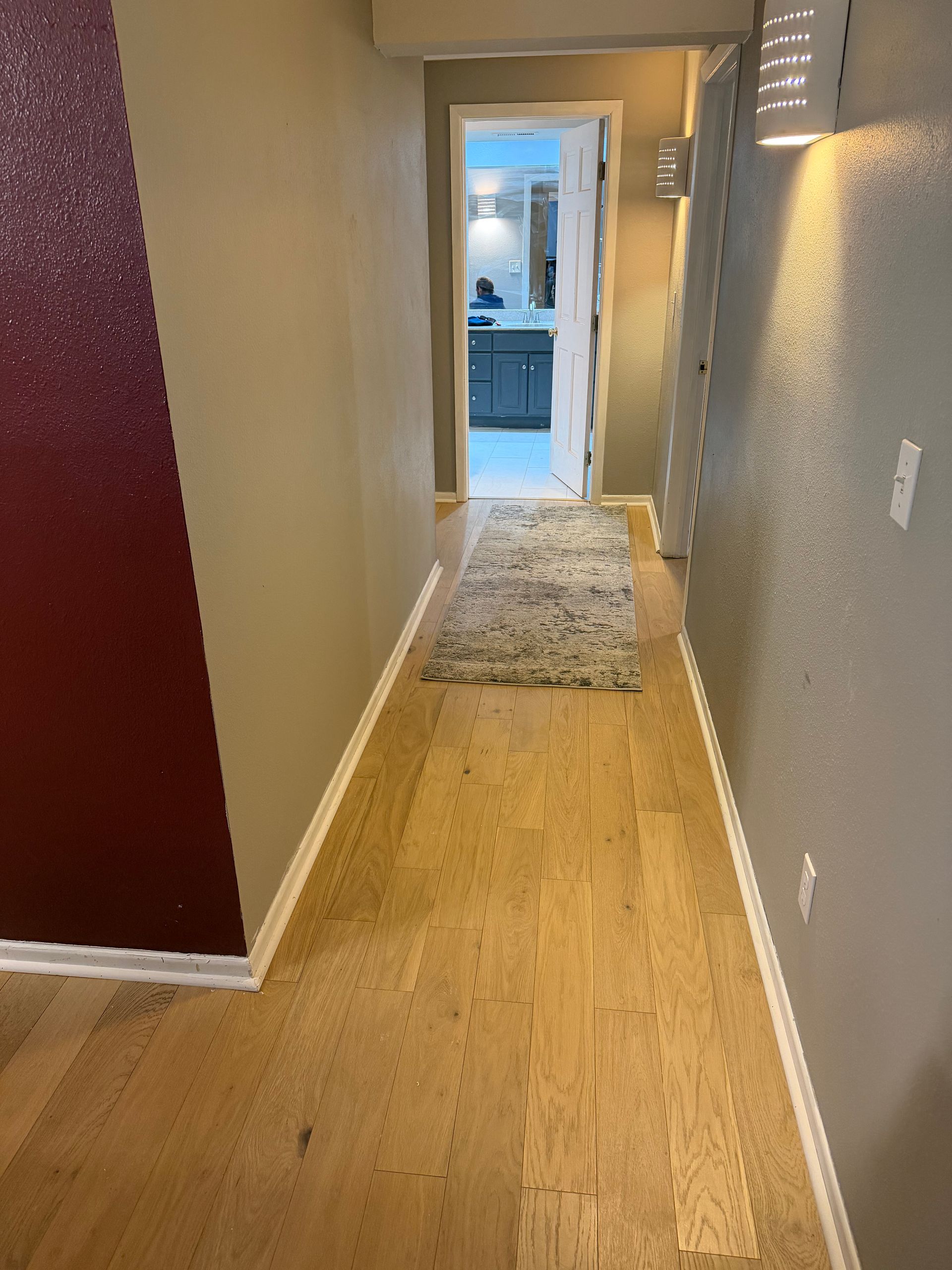 Narrow hallway with light wood floor, beige walls, and a rug leading to a kitchen doorway.