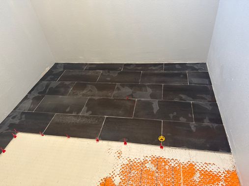 Partially tiled floor in a small room, with unfinished concrete and orange debris in the foreground