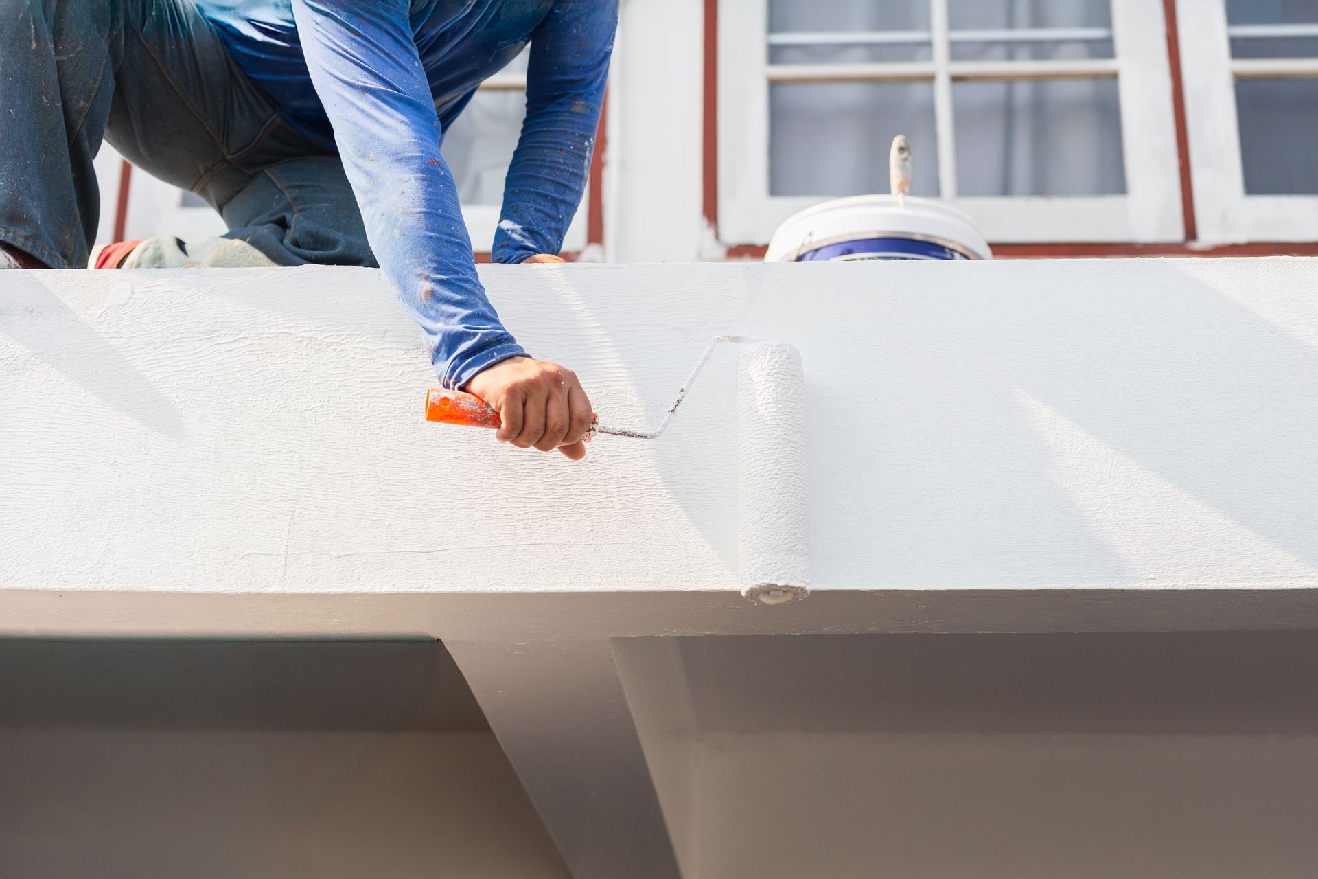 Worker painting a white exterior wall beneath a window with a paint roller