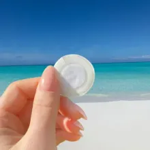 Hand holding a white contact lens case on a bright beach with turquoise water and blue sky