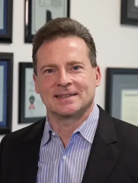 Portrait of a man in a dark blazer and striped shirt, smiling in an office with framed certificates behind him