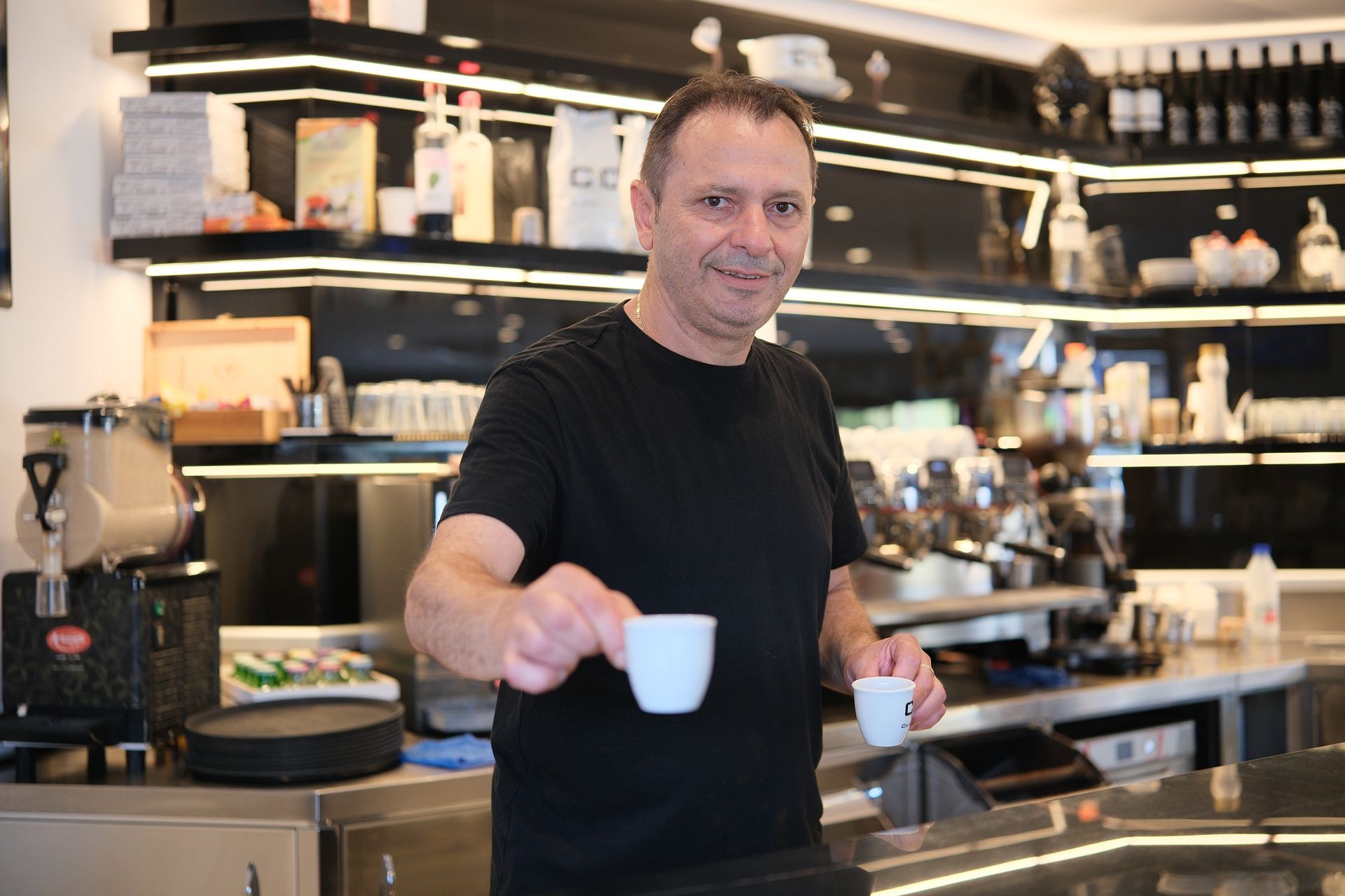 Un uomo con una camicia nera tiene in mano due tazzine di caffè espresso in un bar e sorride.