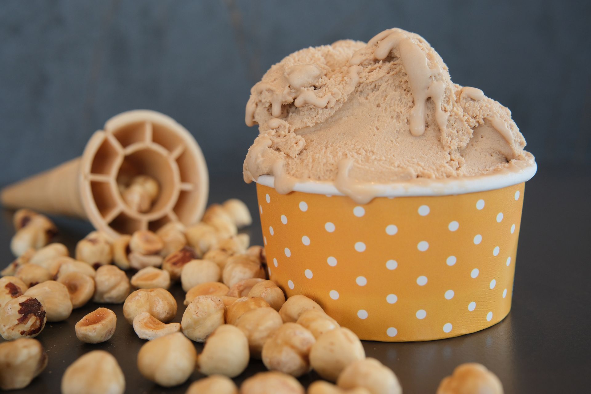 Gelato alla nocciola in una coppetta a pois gialli, cono e nocciole sparse su una superficie scura.