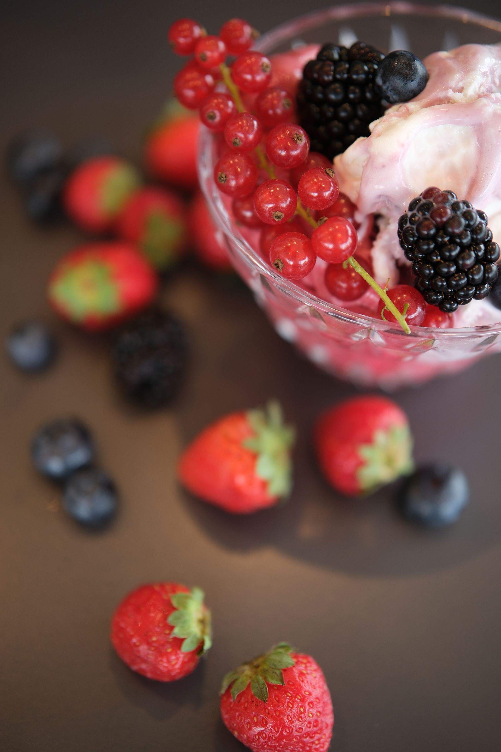 Ciotola di vetro contenente un dessert rosa guarnita con ribes rossi e more, circondata da frutti di bosco sparsi.