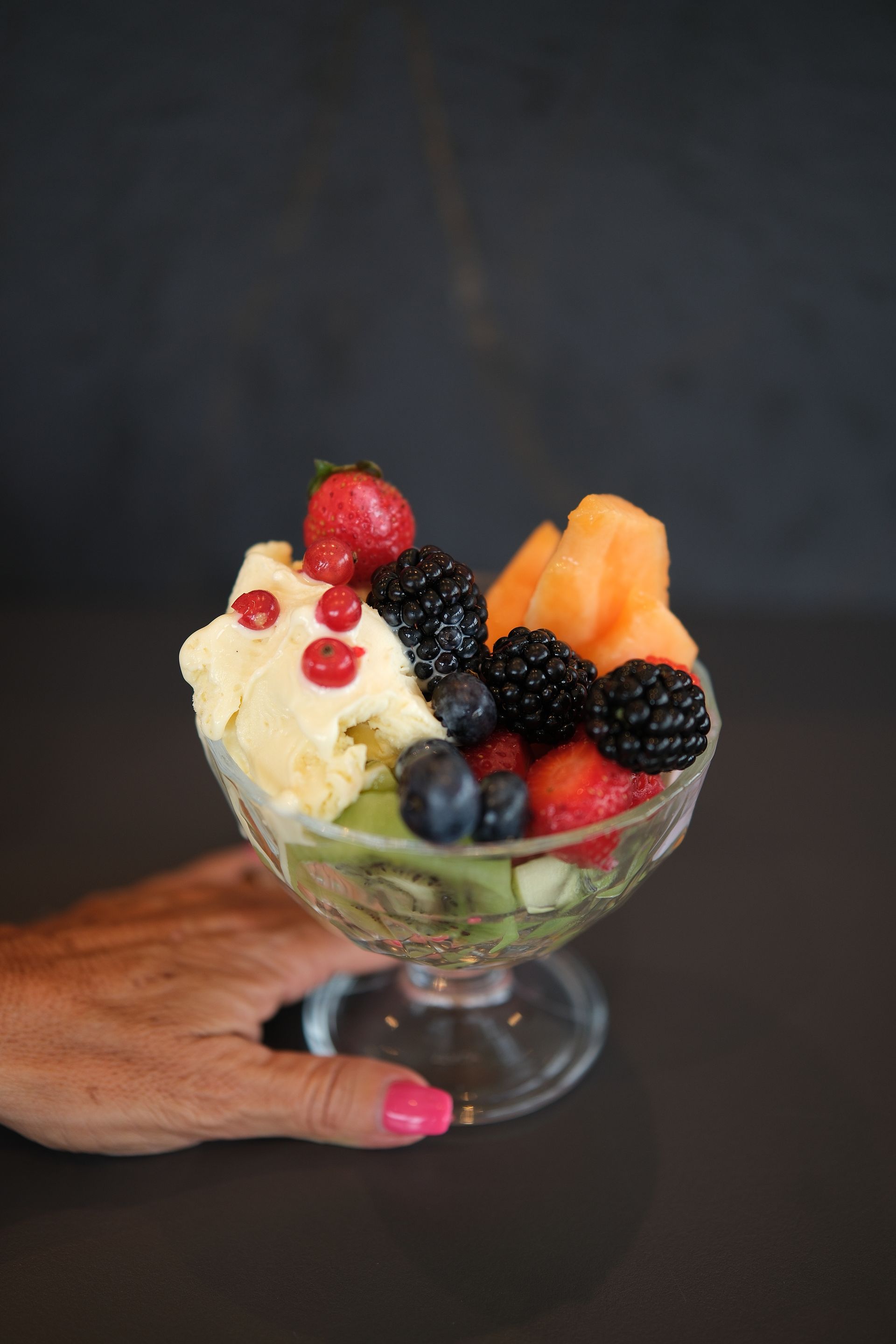 Persona che tiene in mano una ciotola di vetro piena di frutta e gelato su uno sfondo scuro.