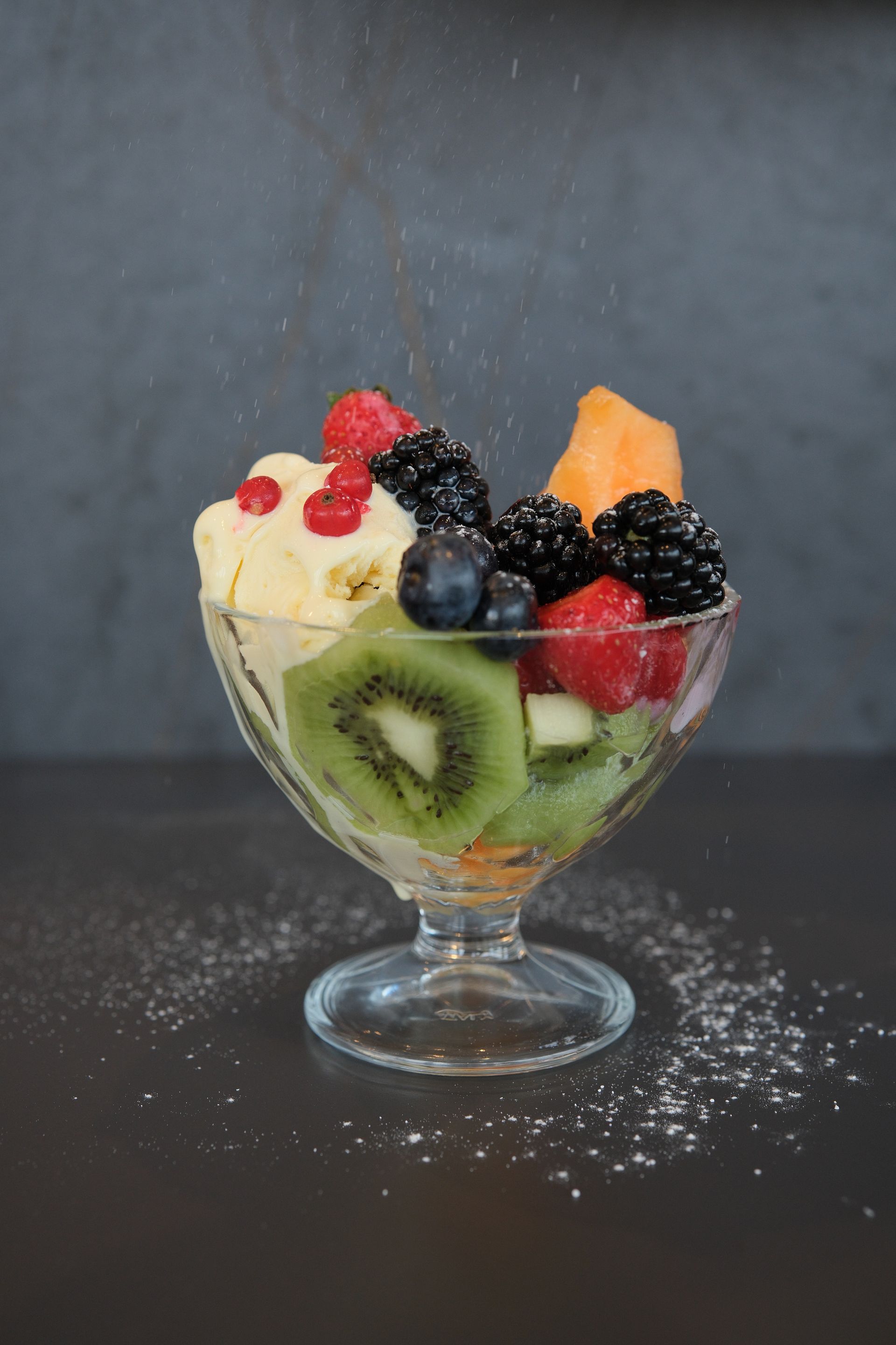 Parfait di frutta in un bicchiere, con kiwi, frutti di bosco, melone e gelato, spolverato con zucchero a velo.