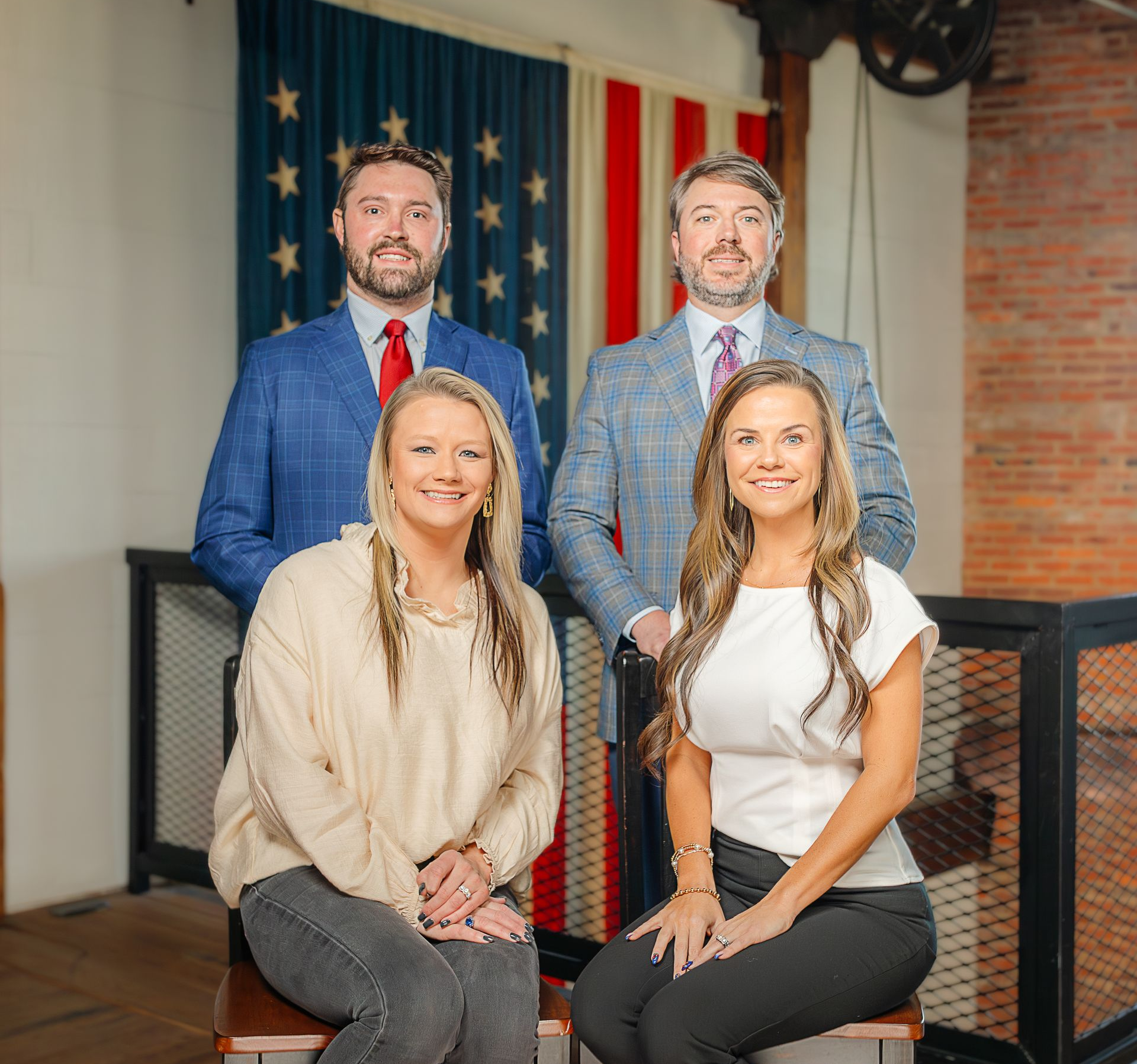 Four people in business attire pose for a portrait in front of a vintage American flag and a brick wall.