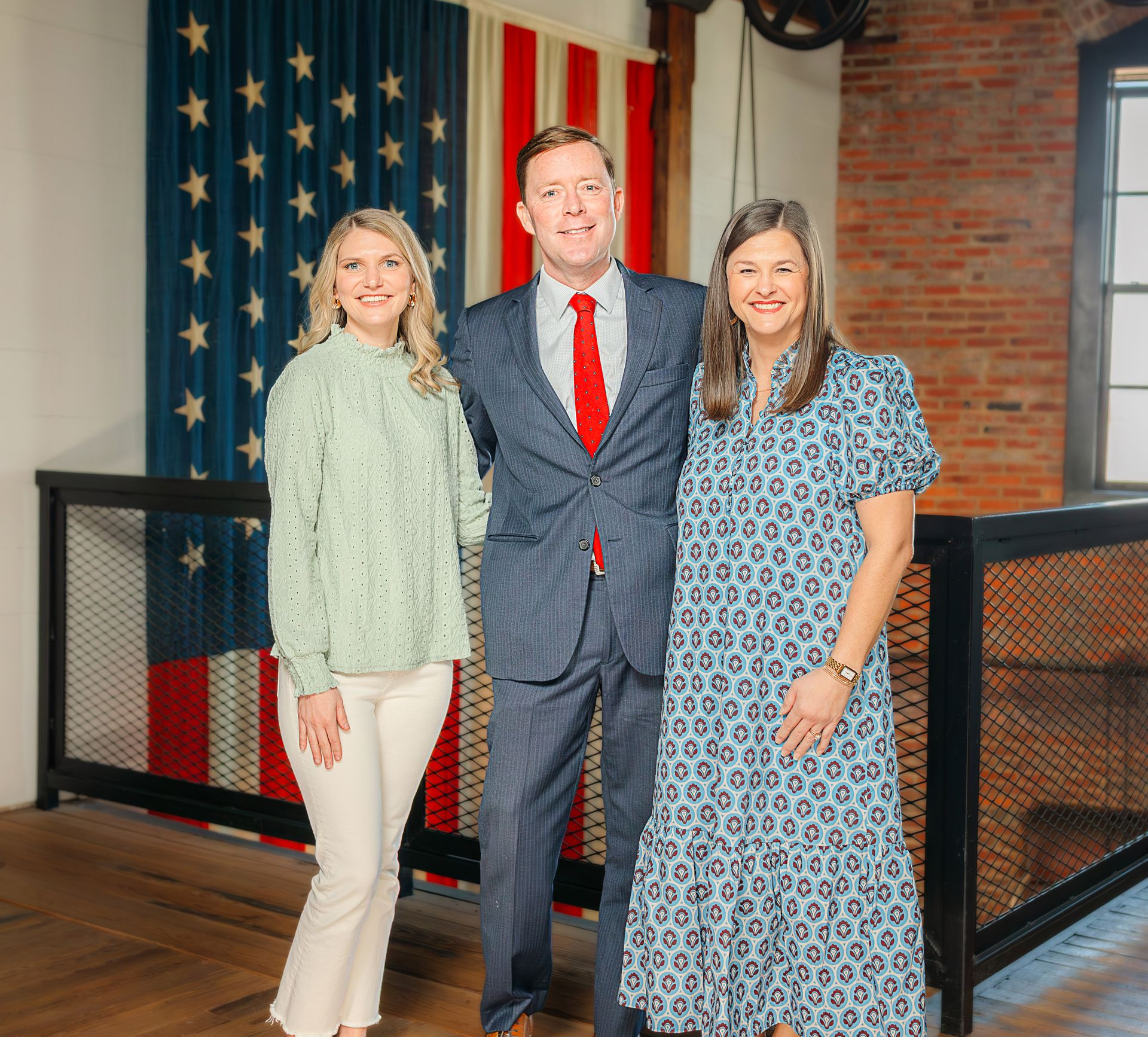 Three people stand smiling in front of an American flag, indoors by a black railing and brick wall.