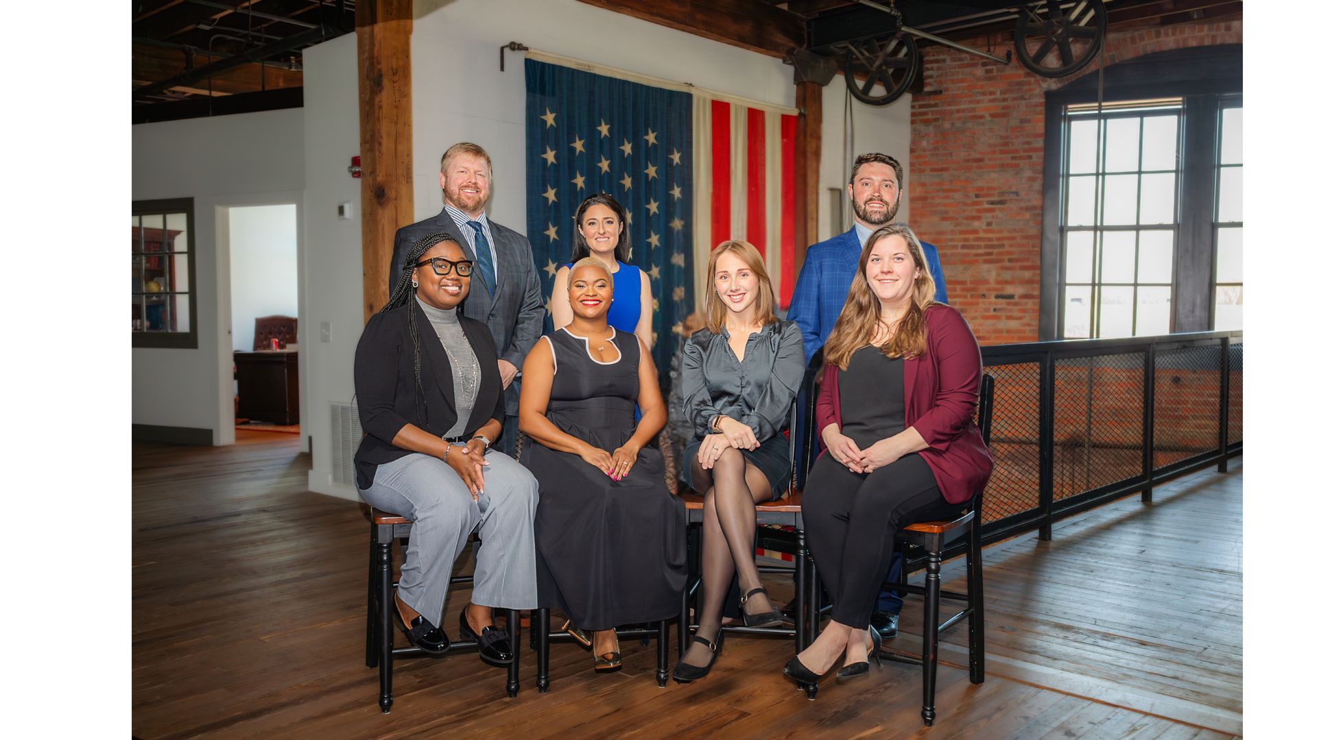 Seven professionals pose in a bright, brick-walled office space in front of an American flag display.
