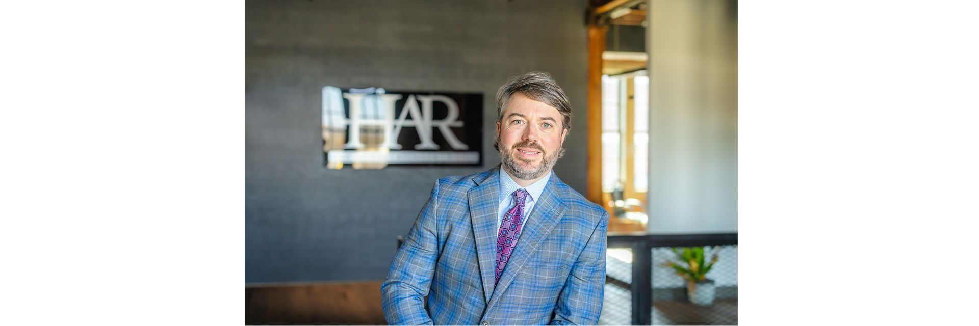 A person in a blue patterned blazer and purple tie stands in an office with an HAR sign on the wall behind them.