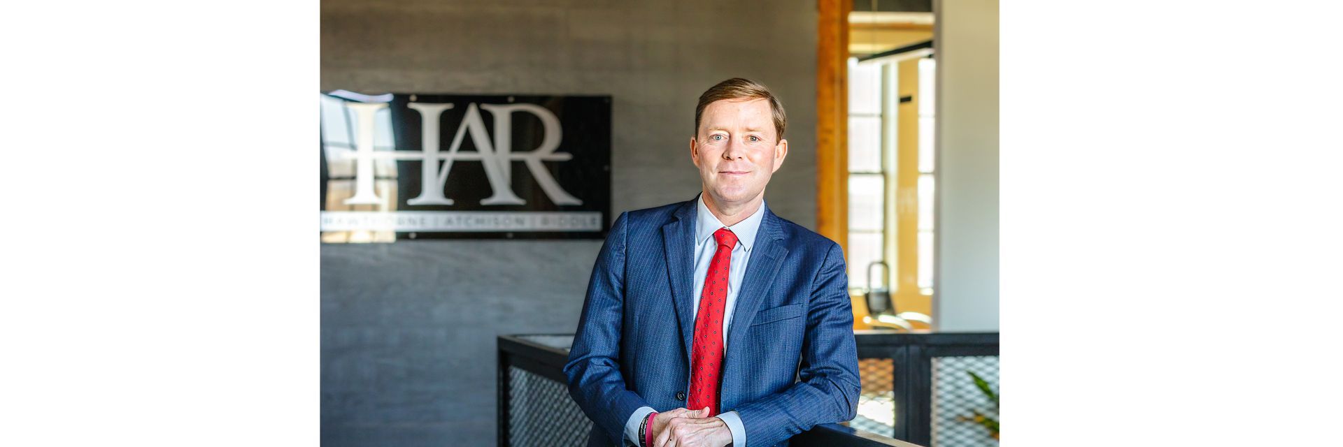 A person in a blue suit and red tie stands in front of a wall featuring a black and silver HAR sign.