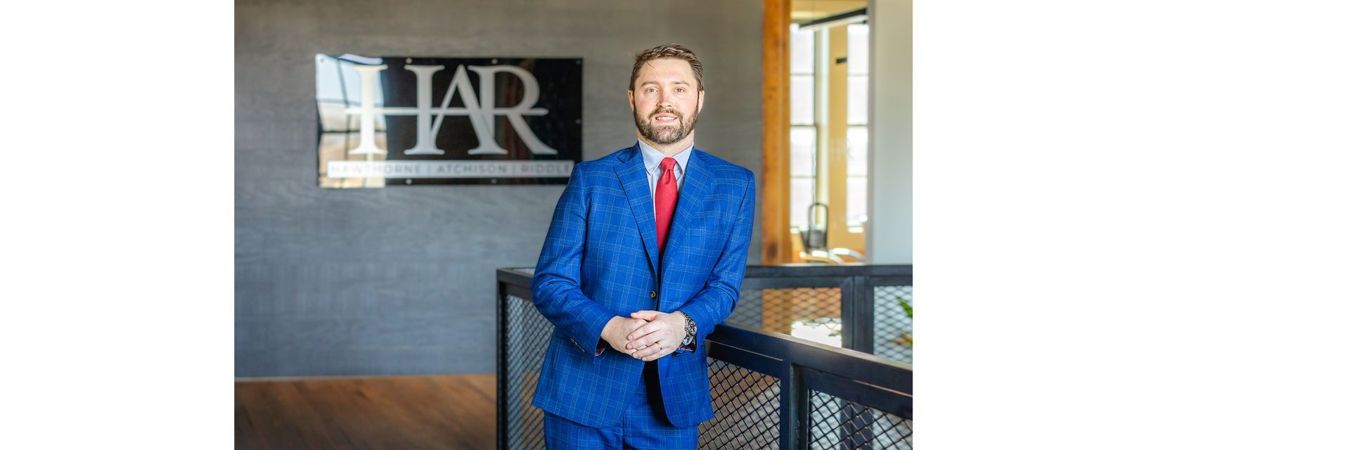 A person wearing a blue plaid suit and a red tie leans against a black railing in an office with a logo on the wall.
