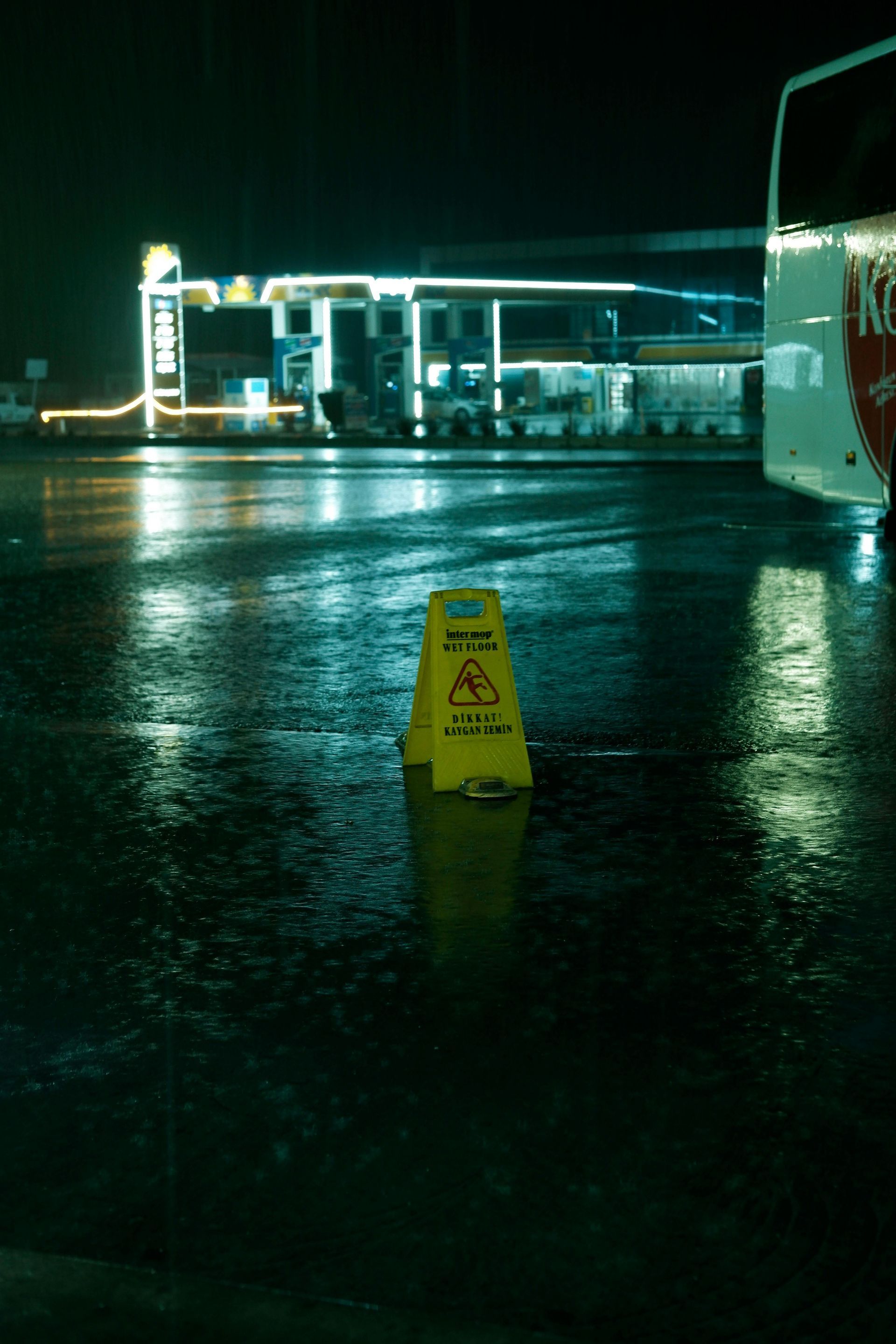 A yellow caution sign stands in a rain-slicked parking lot at night, with a brightly lit gas station in the background.