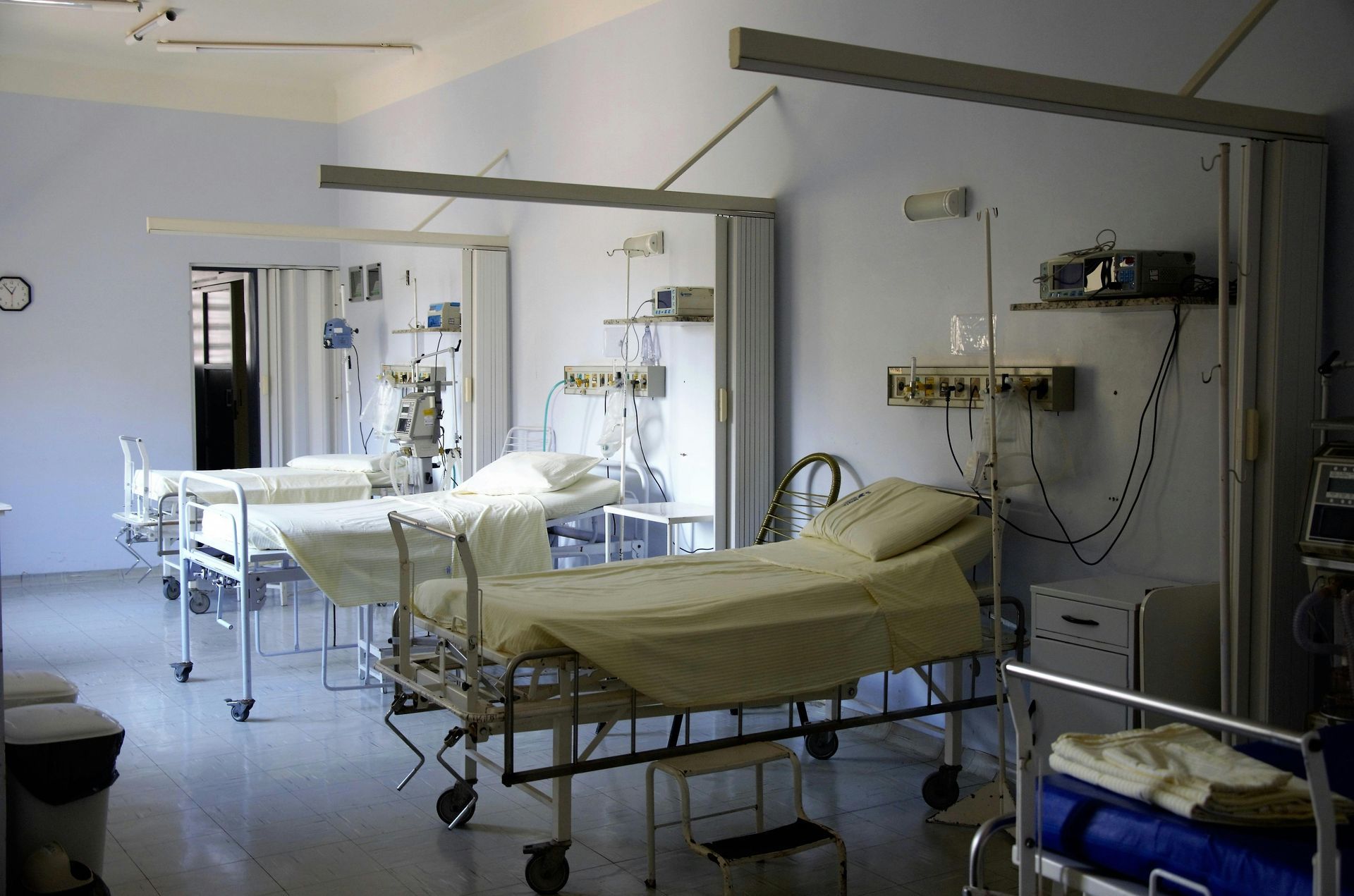 A hospital room with two empty beds, medical monitoring equipment, and curtains suspended from ceiling tracks.