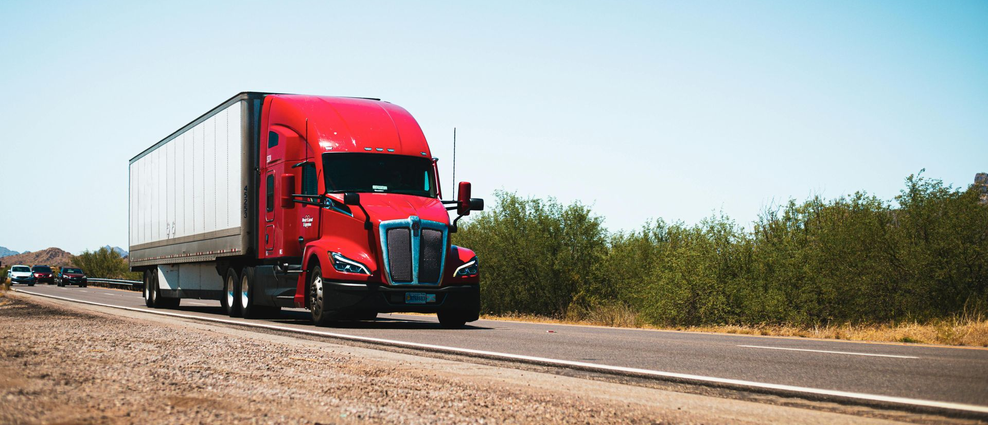 A bright red semi-truck drives down a road lined with green bushes under a clear, bright sky.