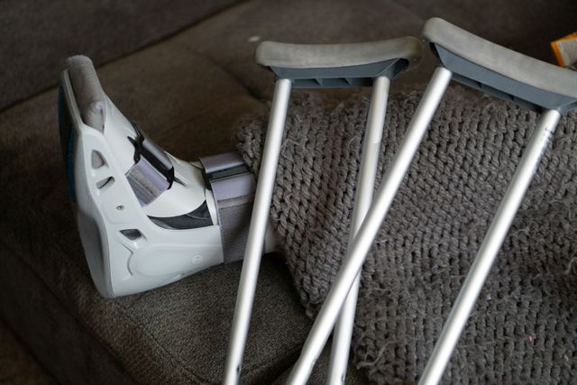 A grey orthopedic walking boot on a leg under a blanket, with two metal crutches resting nearby on a couch.