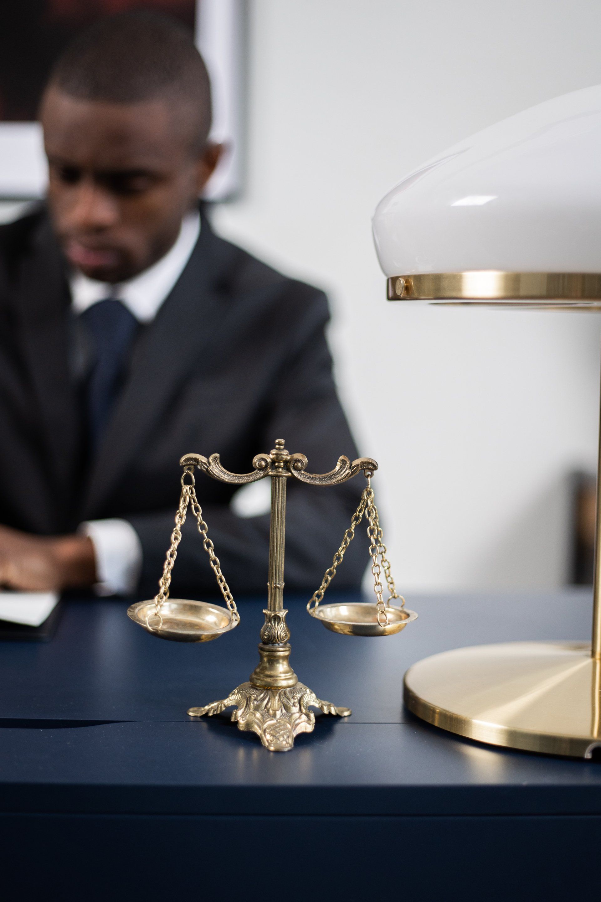 A brass scale of justice sits on a dark desk in front of an out-of-focus professional in a suit and a desk lamp.