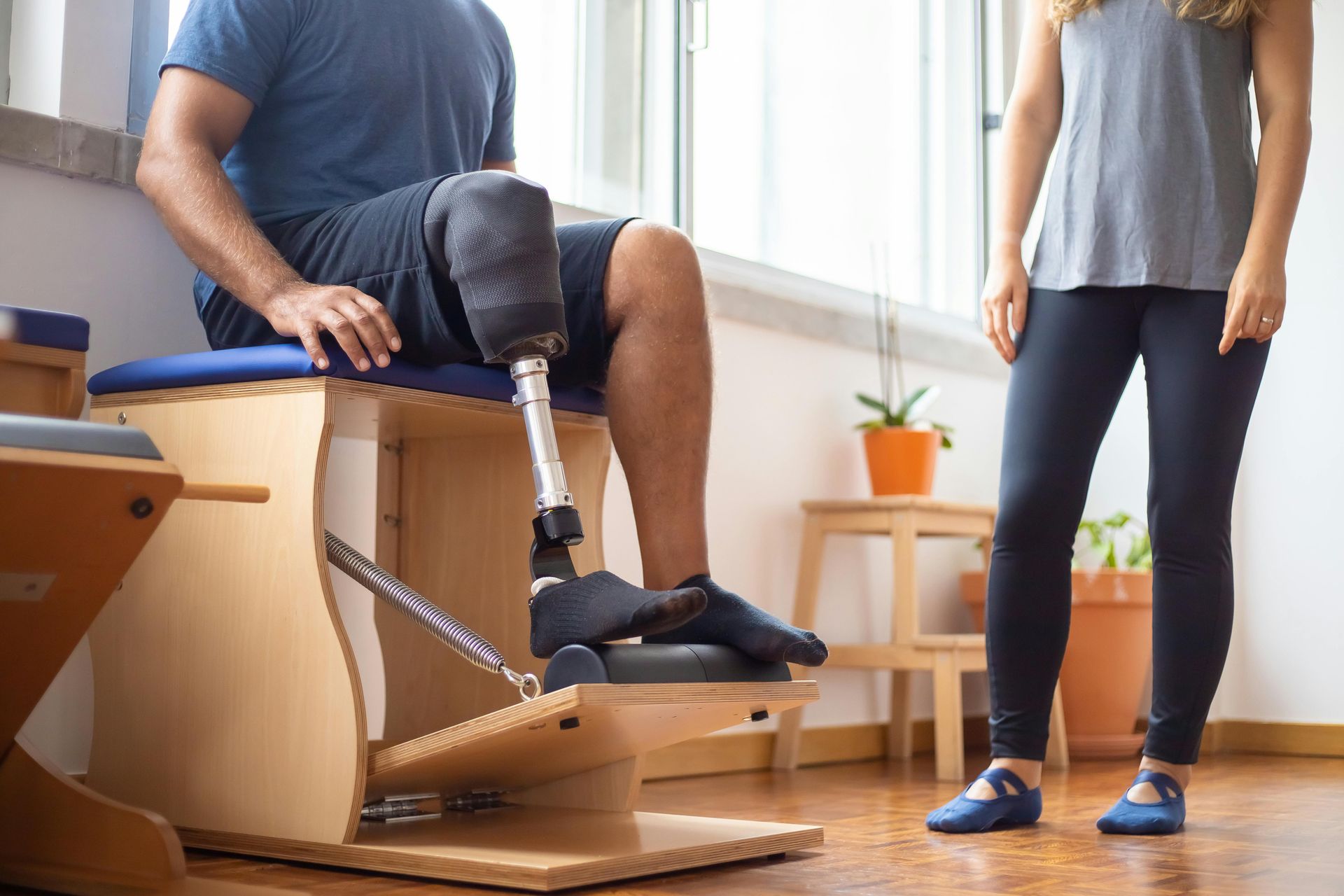 A person with a prosthetic leg uses a Pilates chair for rehabilitation, guided by an instructor in a bright studio.