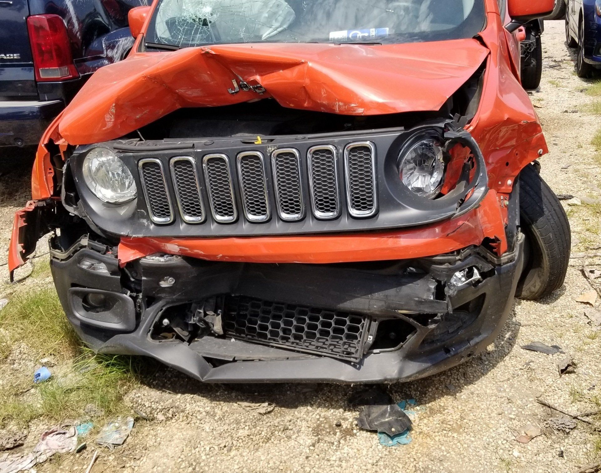 A badly damaged orange Jeep with a smashed hood and front bumper, parked on a gravel lot.