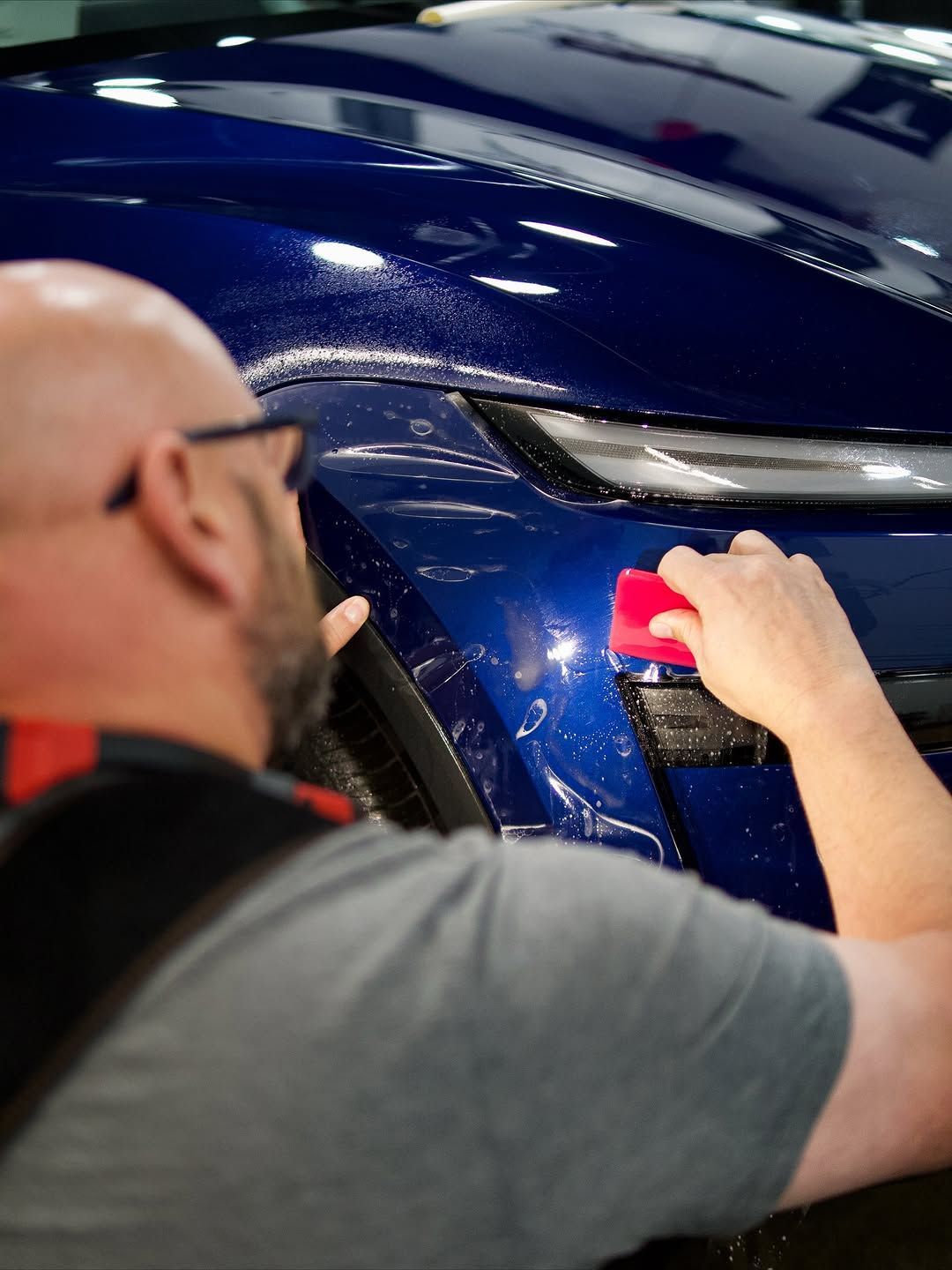 Man applying a blue film to the front bumper of a dark blue car, using a red tool.