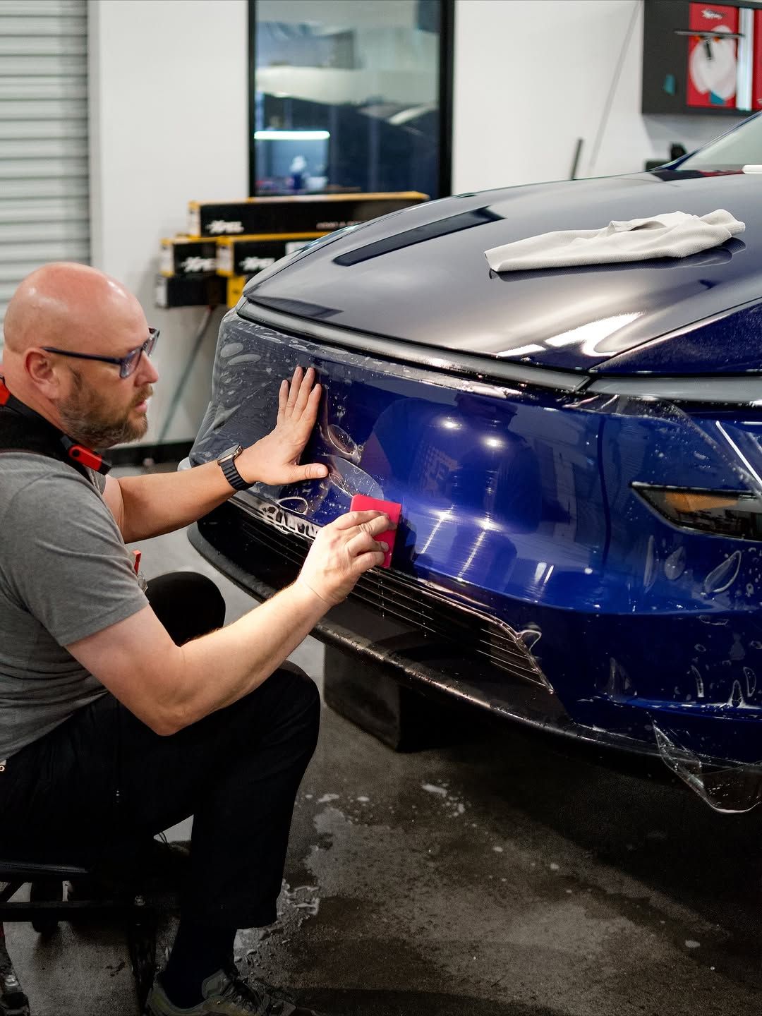 Man applying protective film to a blue car bumper in a workshop setting.