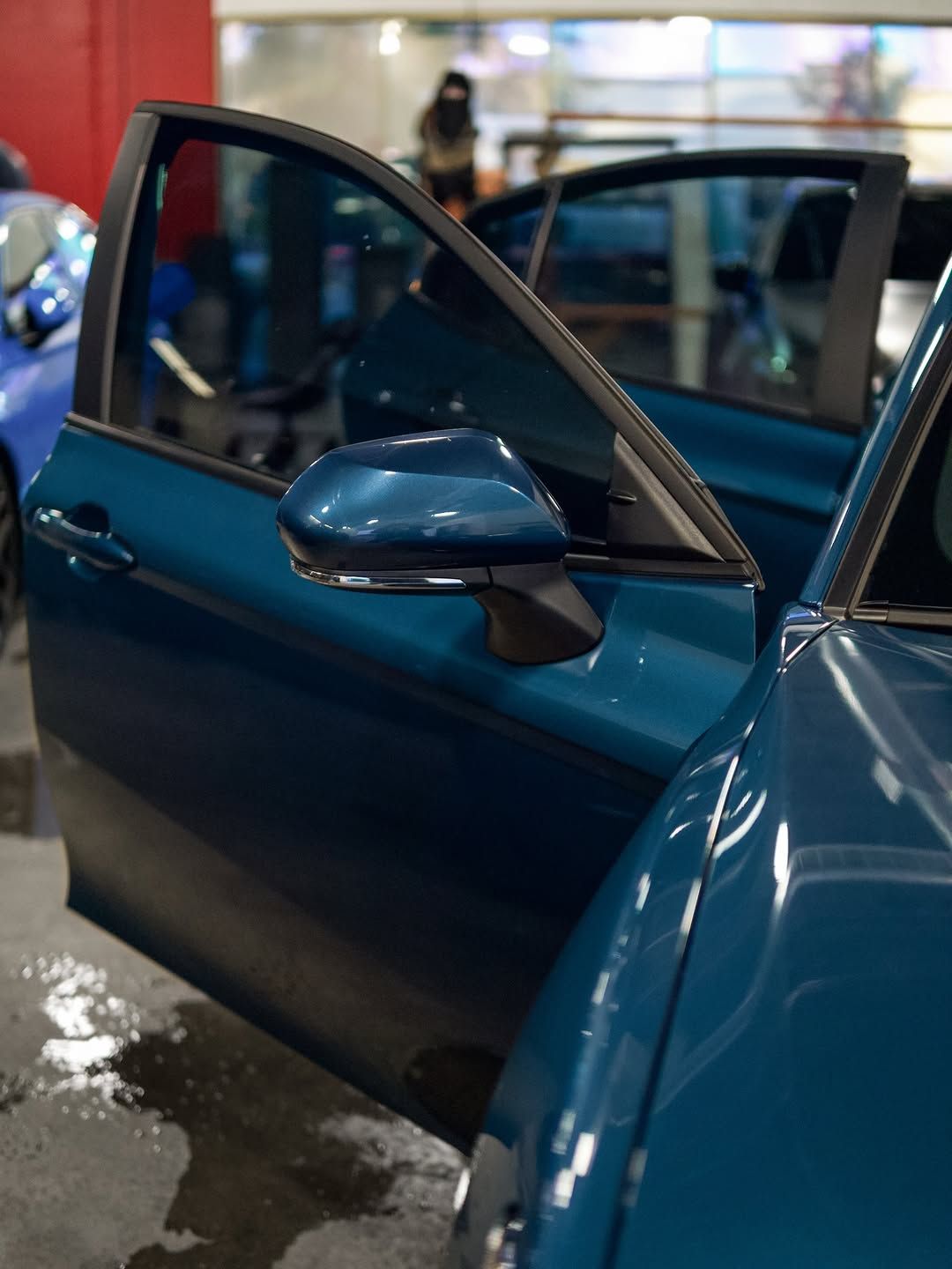 Teal car door open, showing side mirror. Car is in a wet, indoor setting.