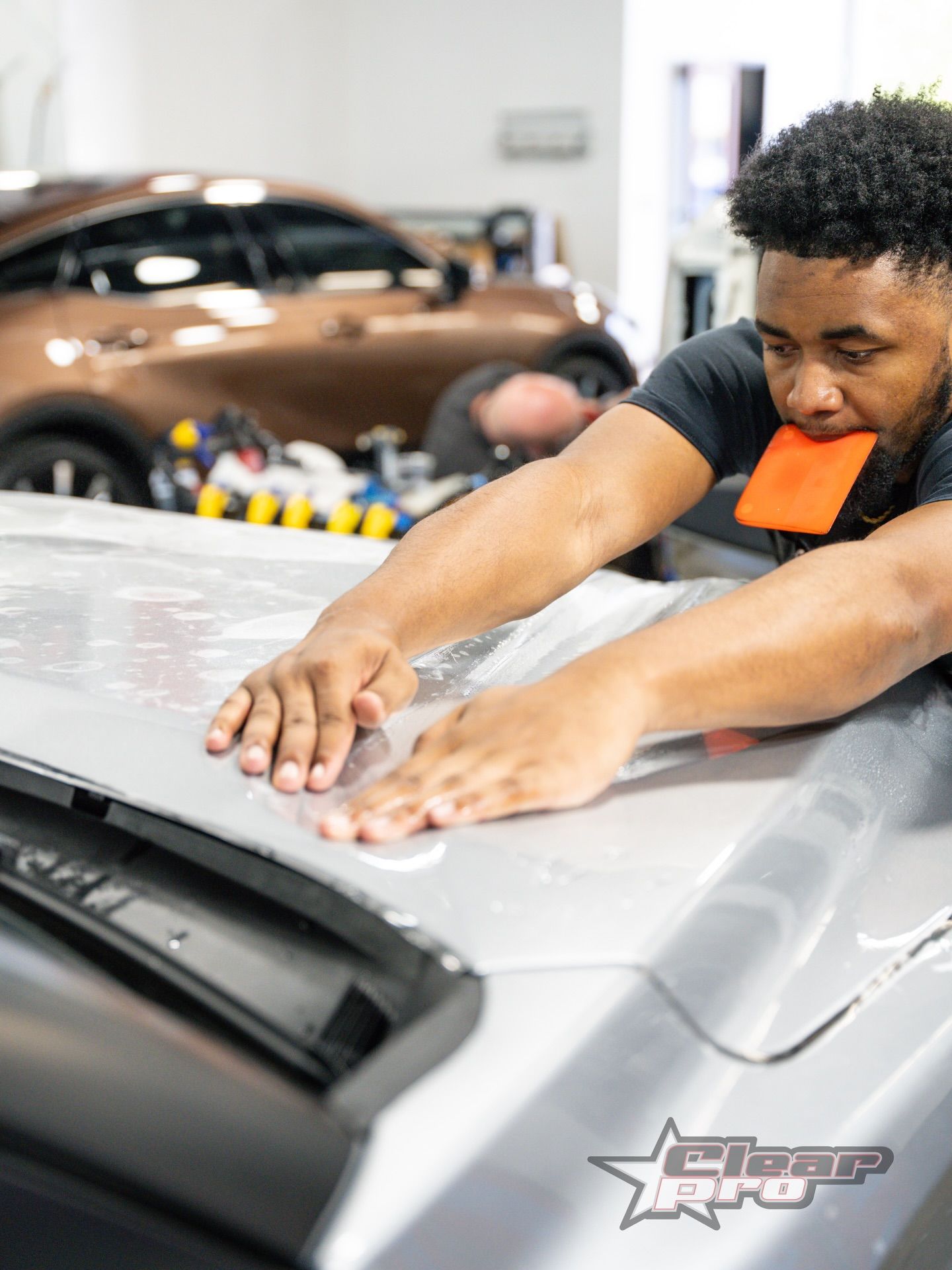 Man polishing a silver car hood in an auto shop with a brown car in the background
