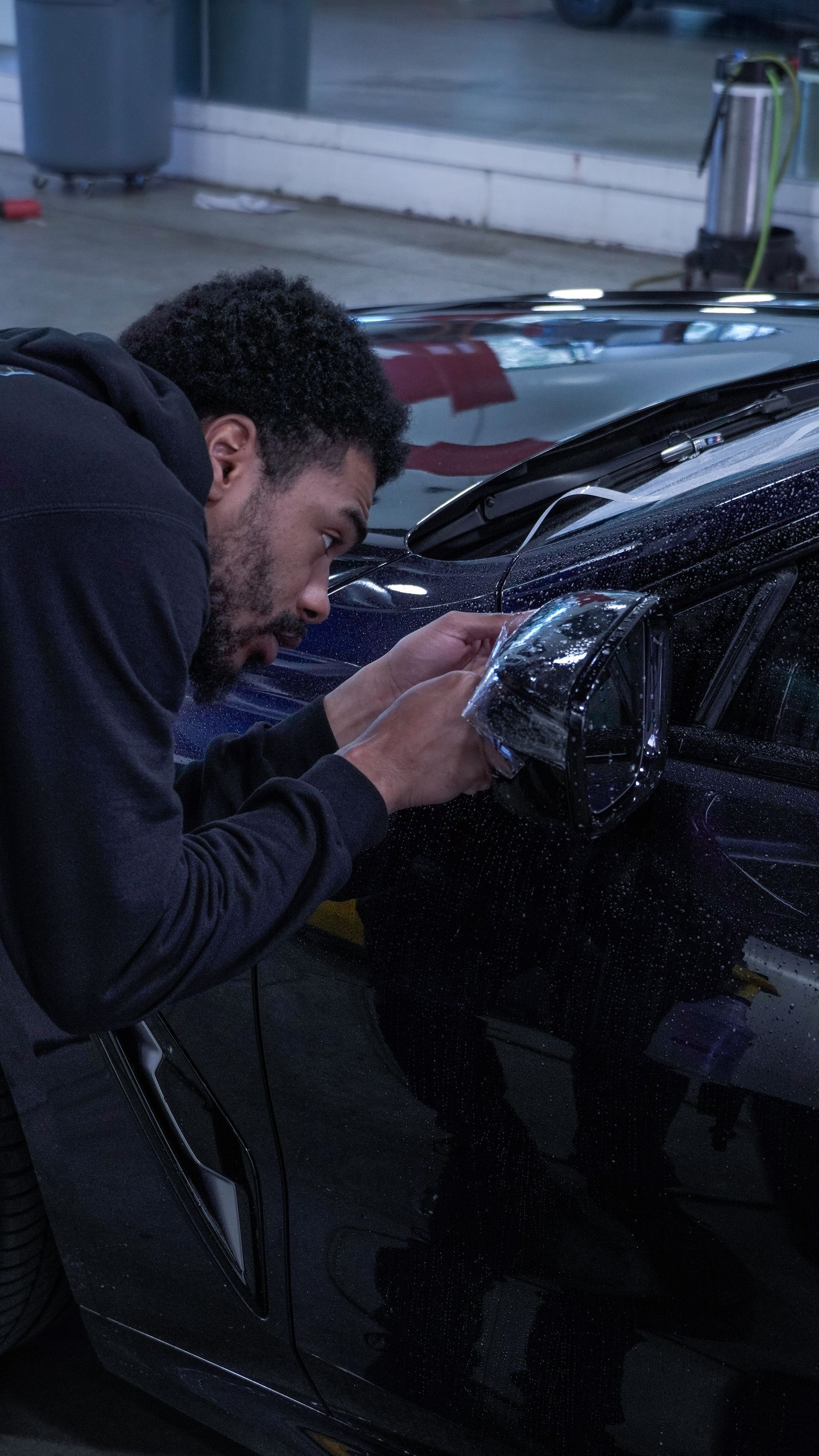 Man applying film to a car's side mirror, in a garage, close up.