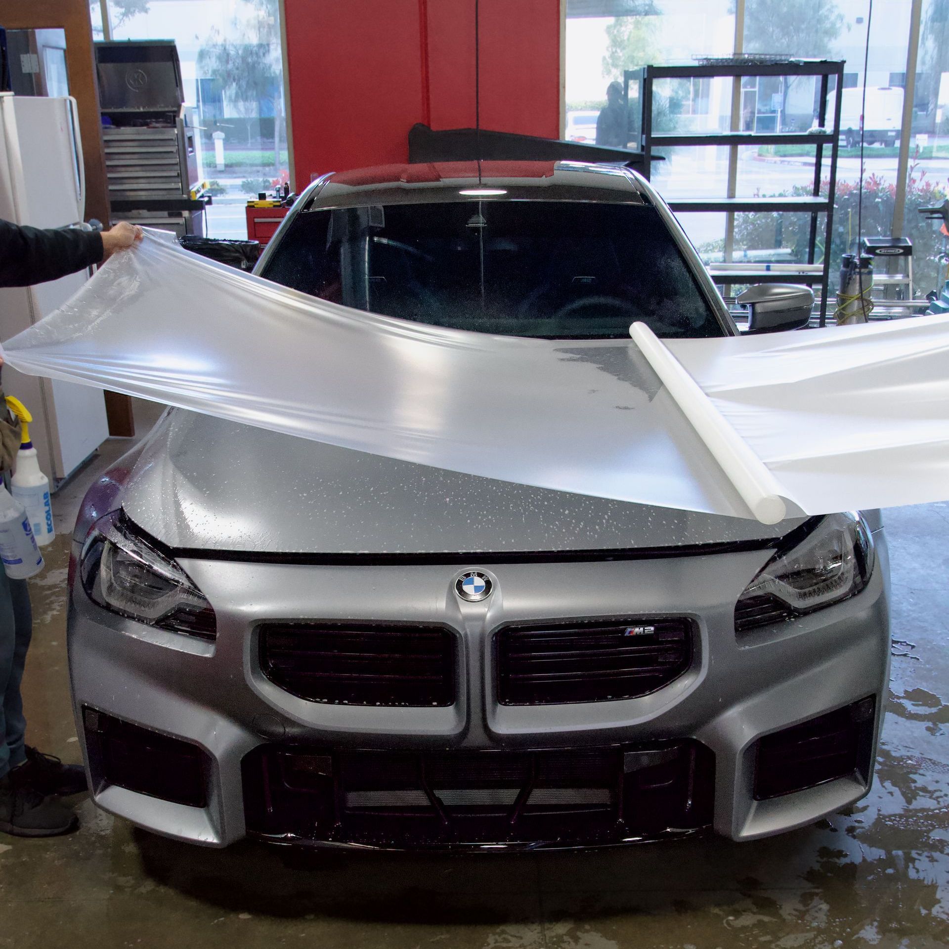 Silver BMW being wrapped in clear protective film, inside a garage.
