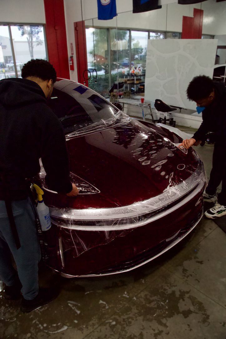 Two people applying film to the front of a maroon car in a shop.