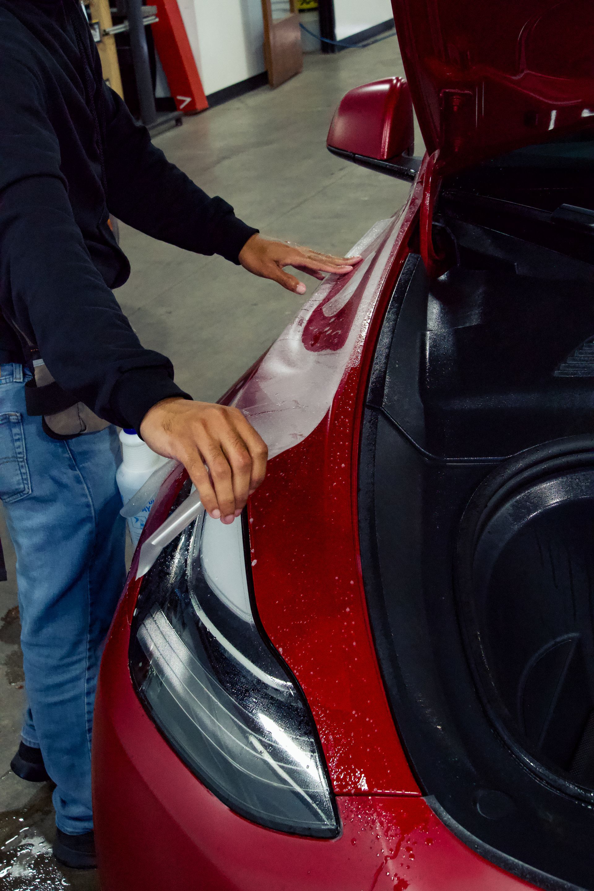 A man is applying a protective film to the hood of a red car.