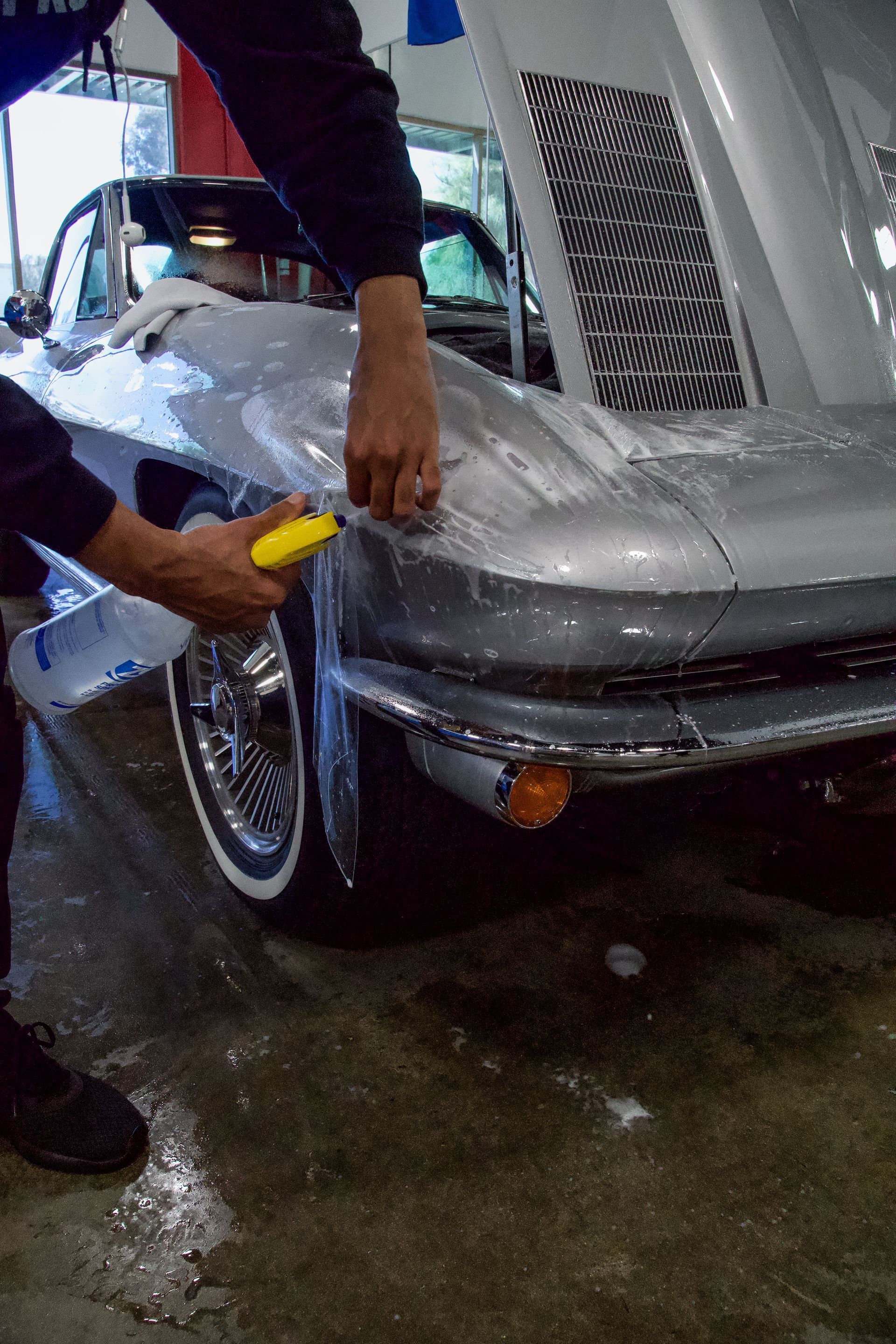 A person cleaning a classic silver car, using a squeegee and a bucket, inside a garage.