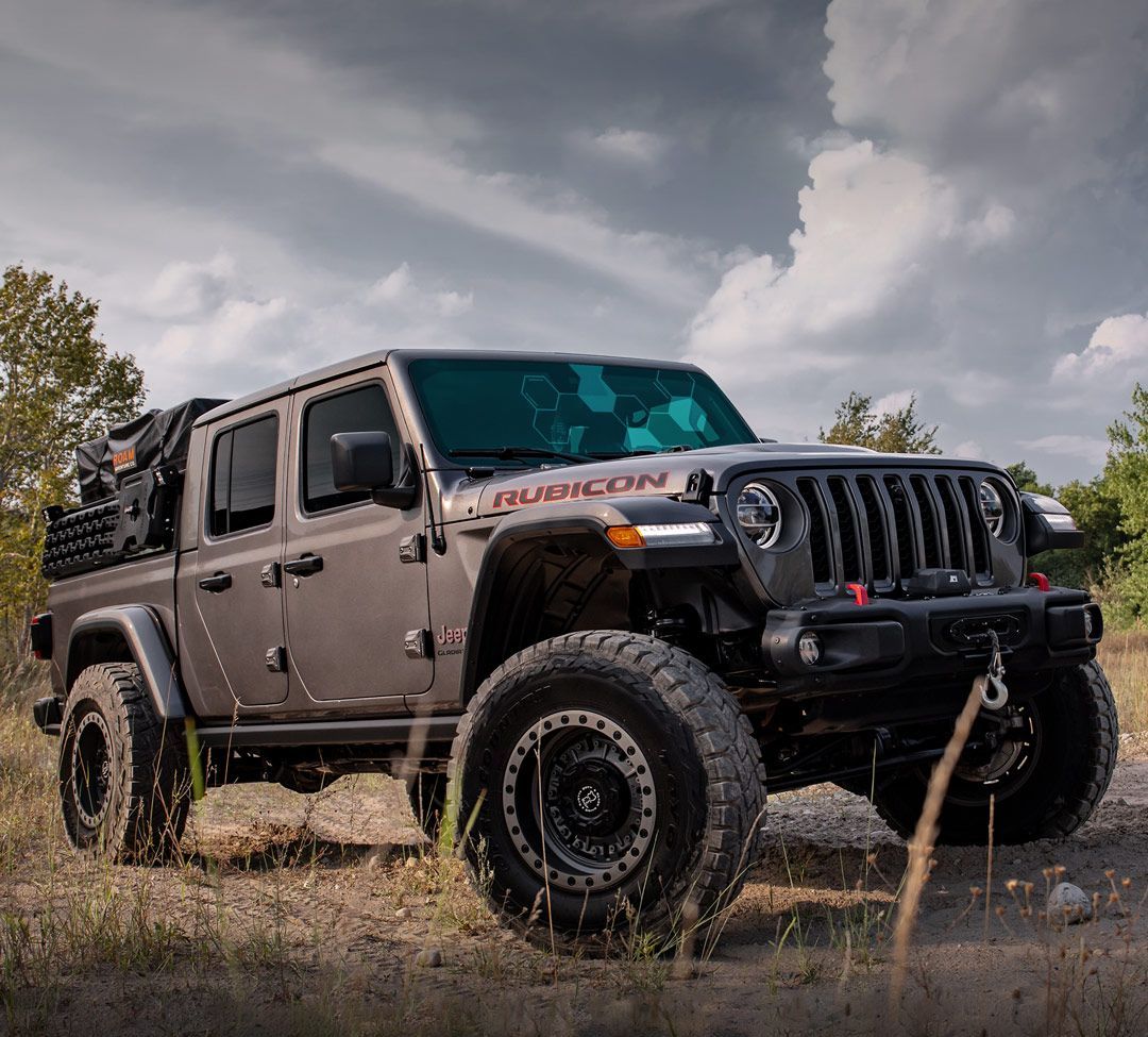 Gray Jeep Gladiator Rubicon off-road truck parked on a dirt path with a cloudy sky background.