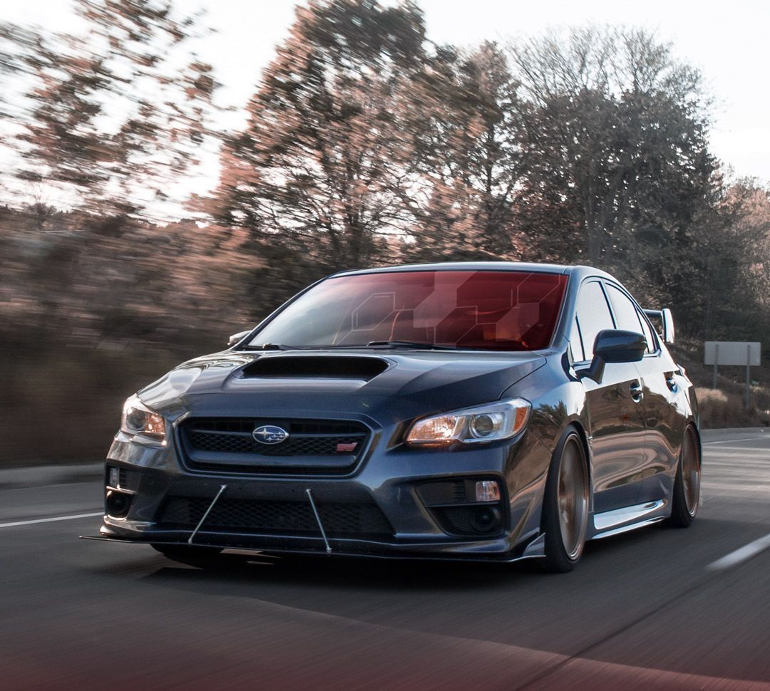 Dark gray Subaru WRX STI driving on a road with a red tinted windshield. Trees are in the background.