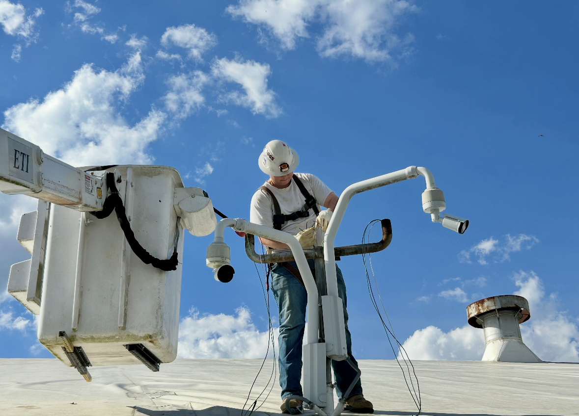 A person kneeling in a grassy field uses a level to install a vertical metal pole into the ground under a blue sky.