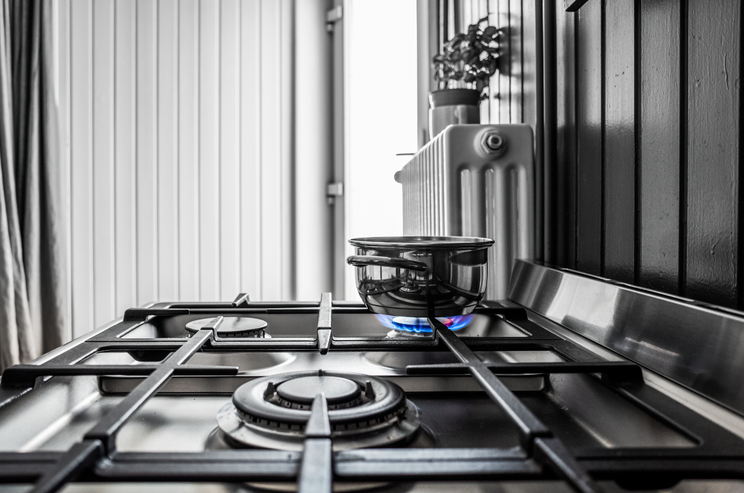 A stainless steel gas stove with a pot on a lit burner, set in front of a window and radiator.