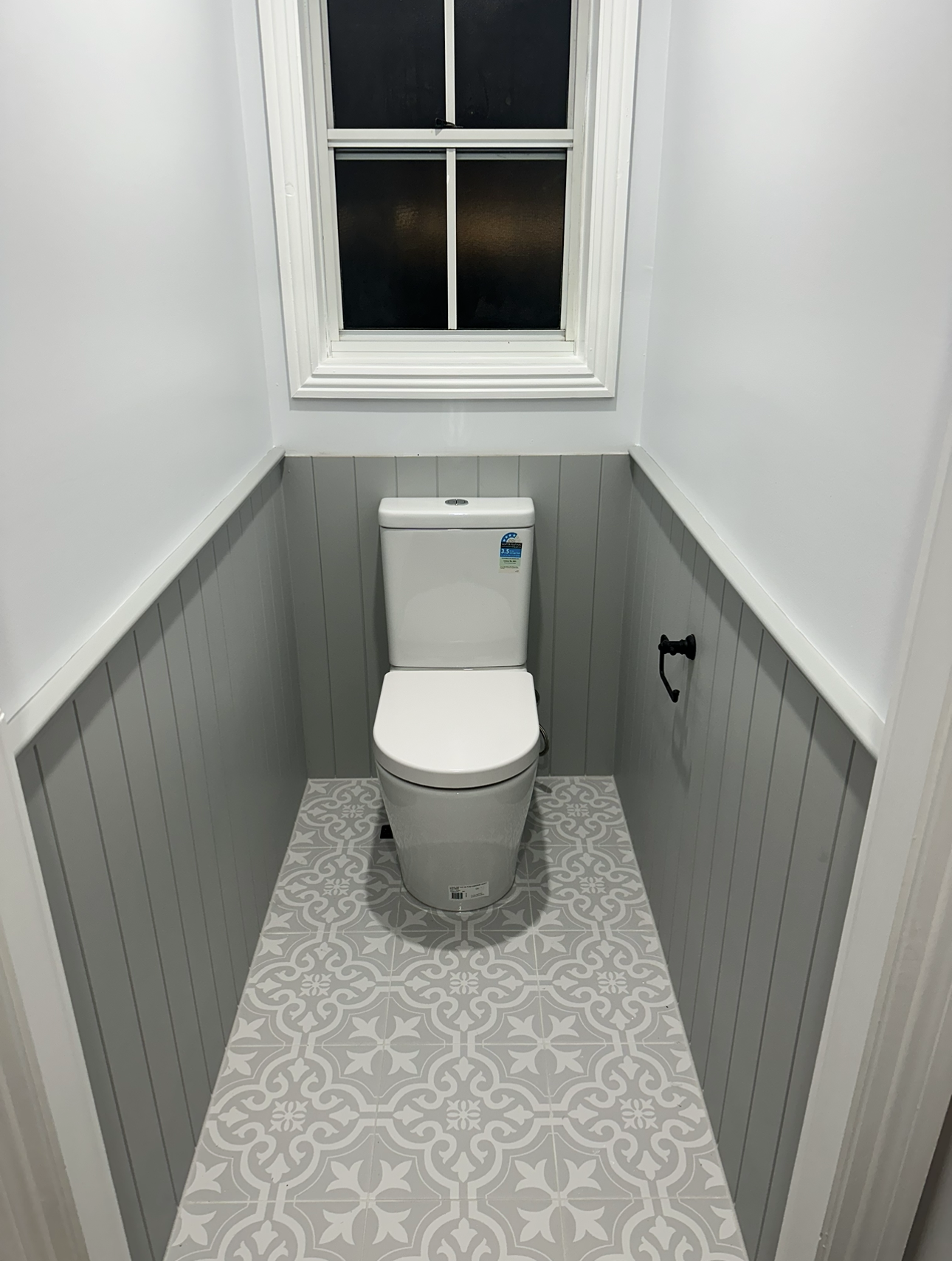 Small, gray and white bathroom with toilet and decorative floor tiles. Window above.