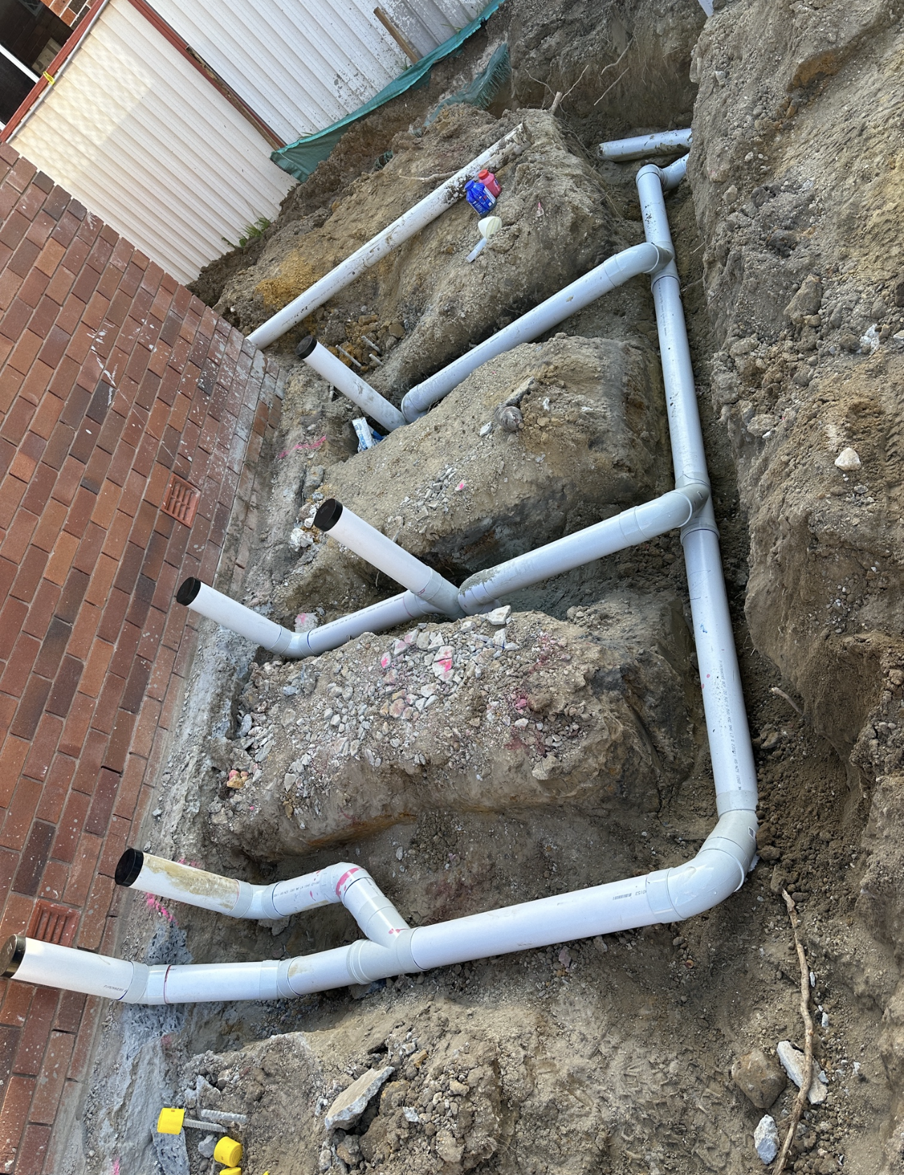 Grey PVC pipes installed in a trench next to a brick wall. Soil is visible.