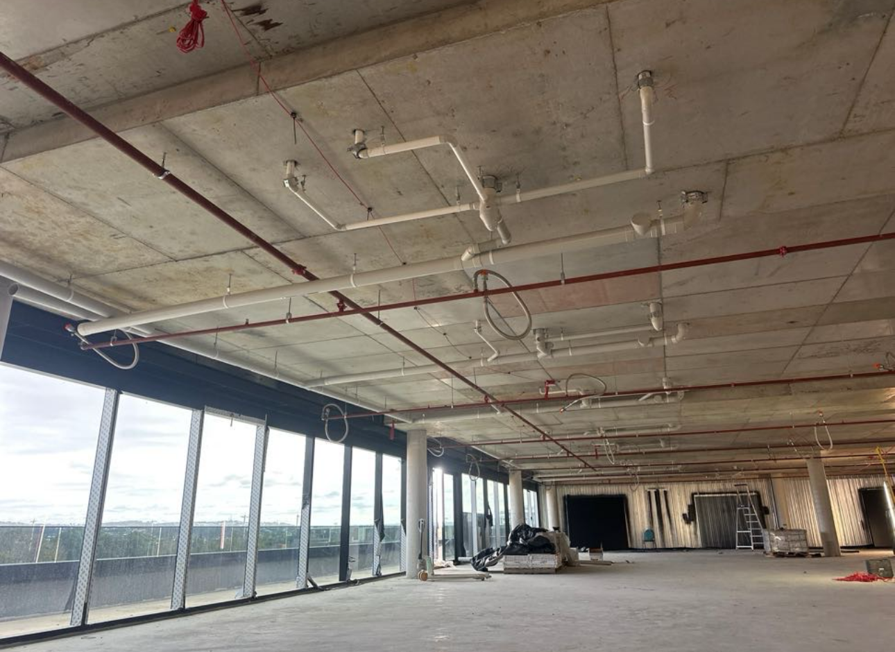 Empty office space under construction, featuring exposed concrete ceiling with pipes and large windows.