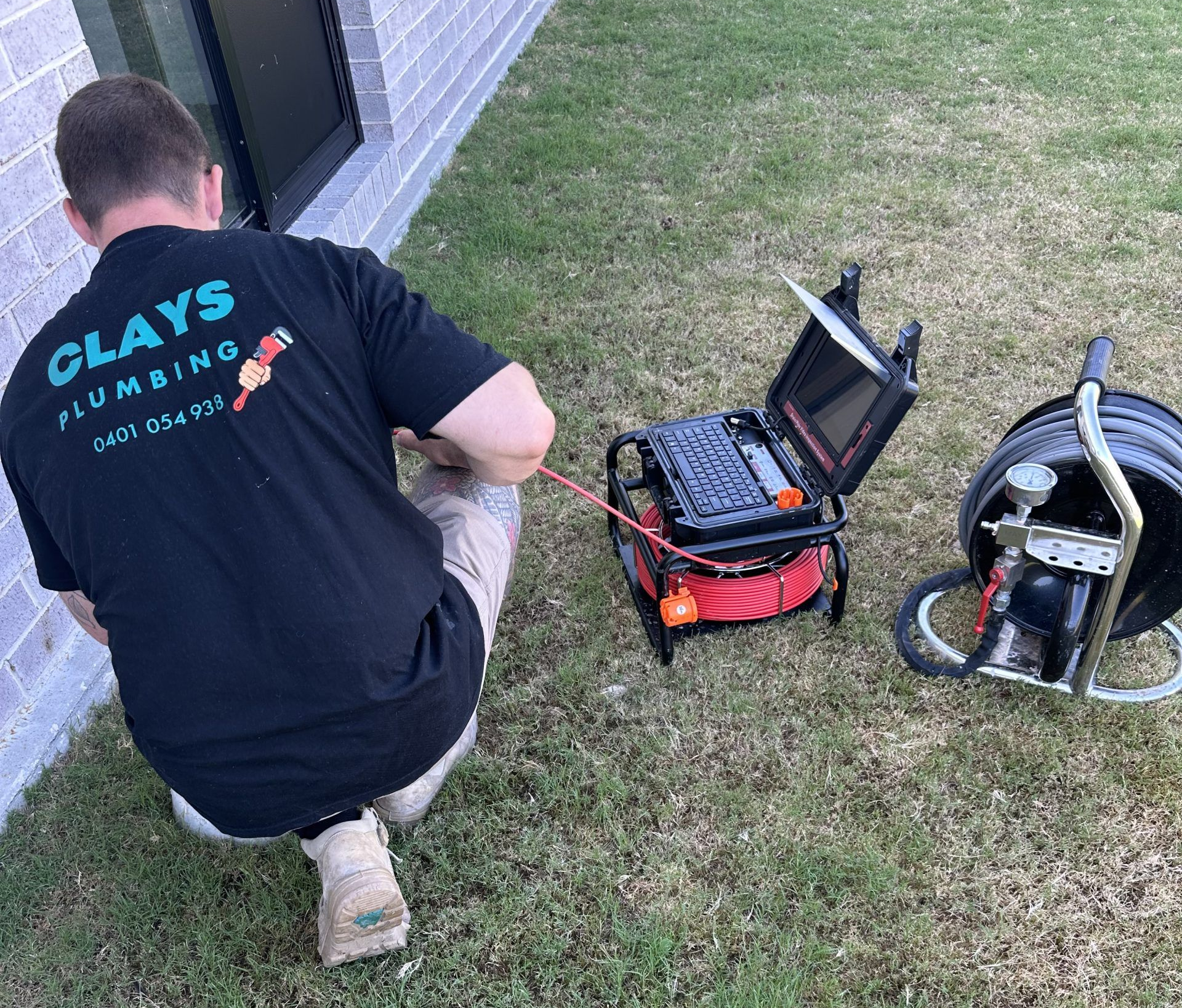 Plumber using a camera to inspect pipes outside a building on grass. Black monitor and reel.